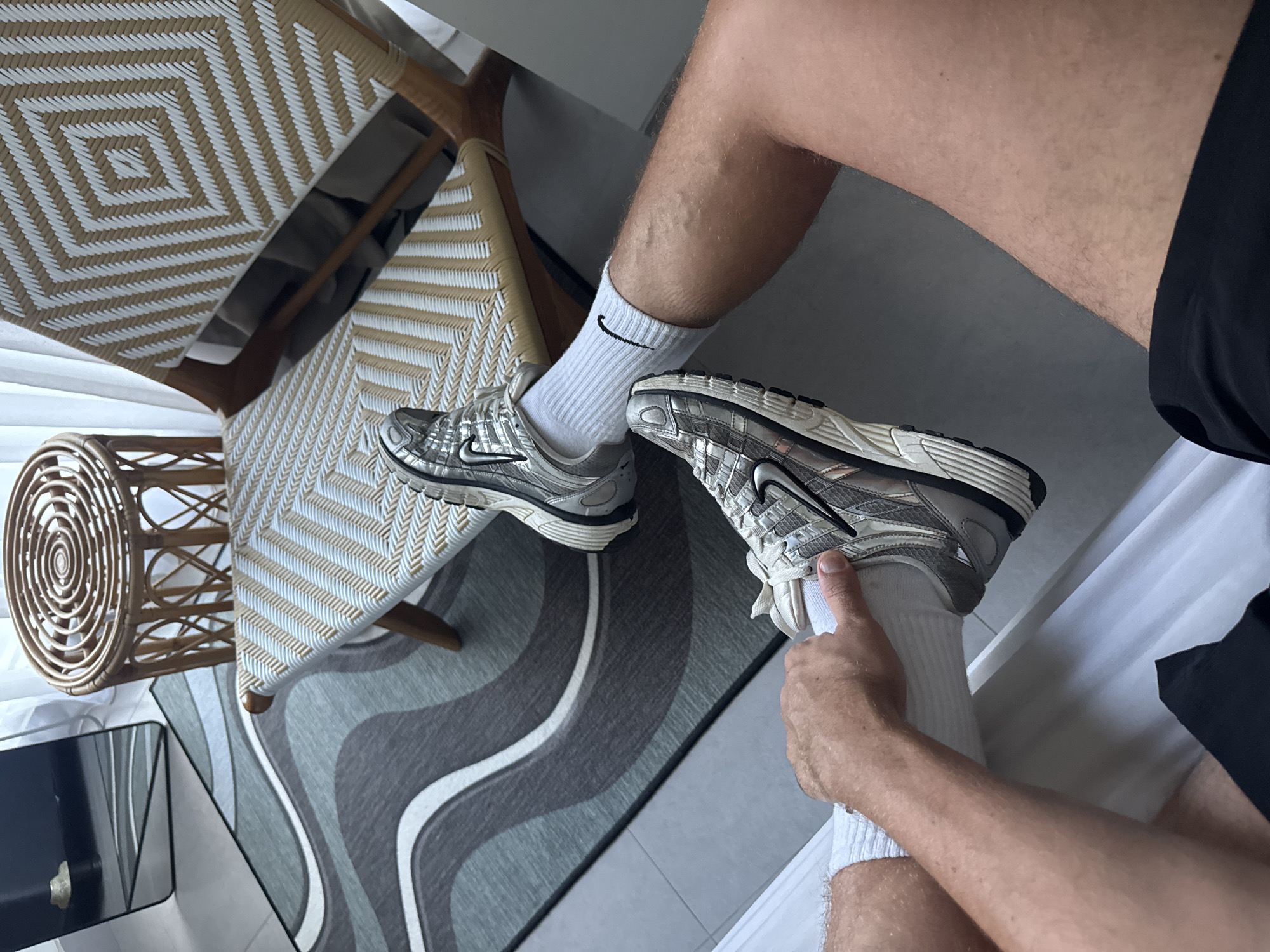 The image shows a first-person perspective of a person sitting in a room, possibly a hotel or residence. The person is wearing a pair of silver Nike running shoes with white socks. The visible parts of the body include legs, arms, and hands. The person appears to be touching the laces of the shoe on the right foot.
The background includes two woven chairs, one with a table attached to it, and a rug with a gray, white, and black swirly pattern. A small round woven table is also visible.
Based on the interior design and the information provided, it is plausible that the location is in North Kuta, Indonesia, although no specific landmarks or details confirm this.