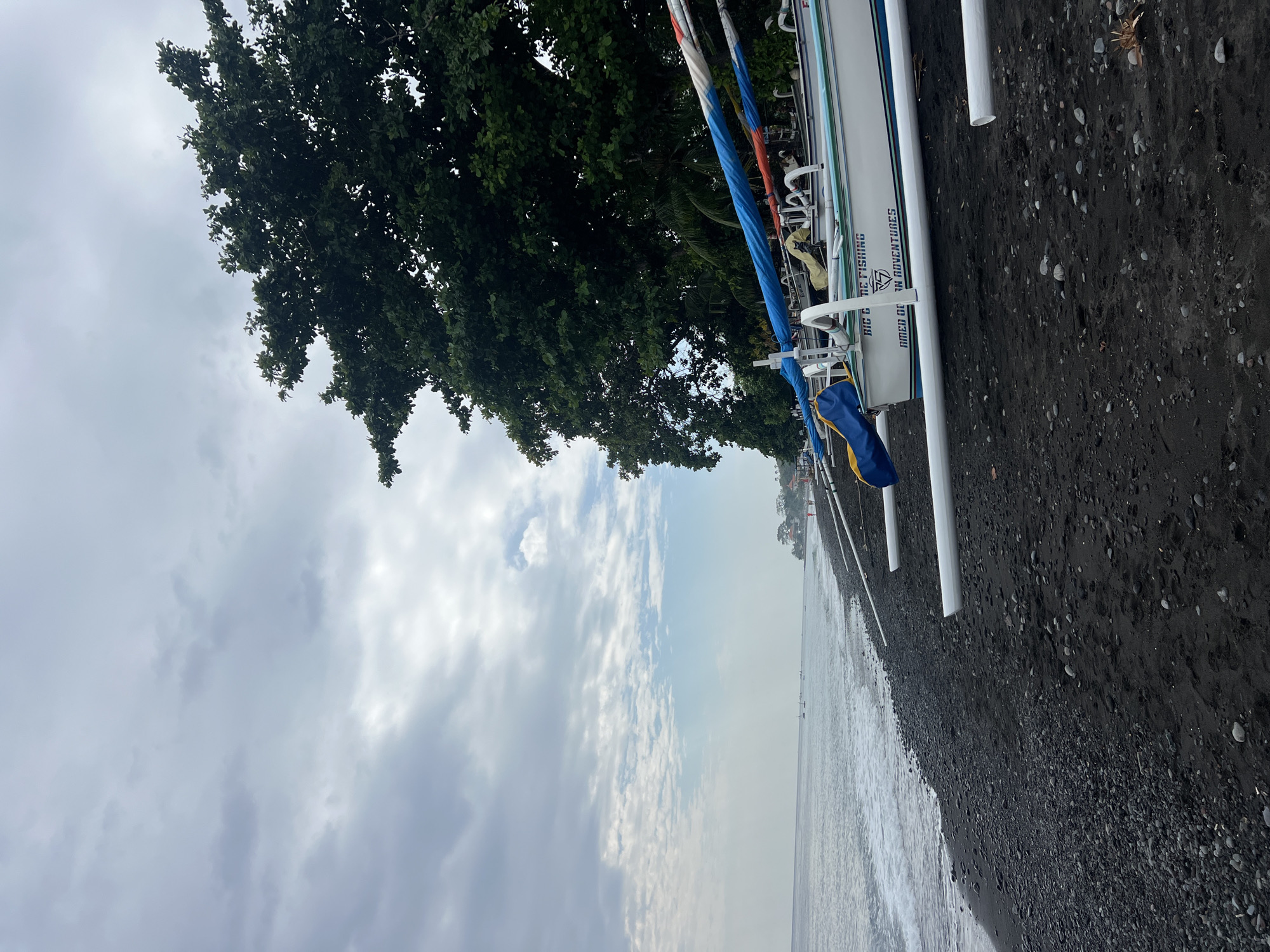The image shows a tranquil scene on a black sand beach in Abang, Indonesia. Several white outrigger boats are lined up on the shore, their colorful canopies a vibrant contrast against the dark sand and muted sky. The gentle waves of the ocean lap at the shore, and in the distance, a few small boats are visible on the water. Lush green trees frame the scene on the right, their dense foliage suggesting a tropical environment. The sky is overcast, with a mix of grey and white clouds creating a soft, diffused light. The overall impression is one of peacefulness and natural beauty.