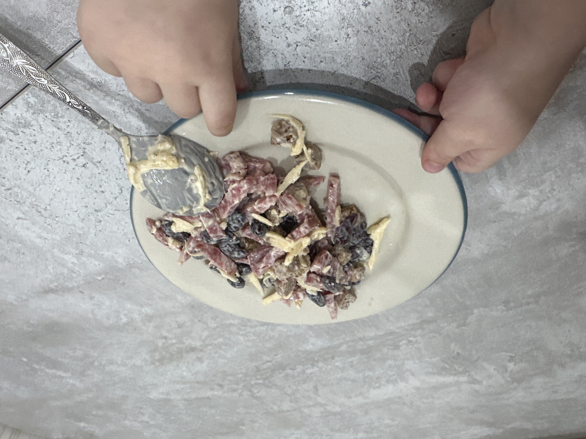 An overhead shot captures a pair of hands, likely a child's, holding a plate of food on a light grey, mottled surface resembling a countertop. The left hand holds an ornate silver spoon, partially coated with creamy dressing and food, hovering over the plate. The right hand supports the edge of the oval, off-white plate, which has a thin blue rim.
The plate contains a mixture of ingredients, resembling a salad. Visible components include dark, possibly dried, fruit or berries (like prunes or raisins), thin strips or shreds of a light-colored ingredient (potentially cheese or vegetable), and reddish-pink pieces of what appears to be cured meat, such as salami or ham. All ingredients are coated in a creamy white dressing, possibly mayonnaise-based, suggesting it's a prepared dish rather than individual components.
The activity appears to be eating or preparing to eat, with the spoon poised to scoop a portion of the mixture. The setting is indoors, likely a kitchen or dining area. There are no visible texts, location-specific cues beyond the general context of a home, or indicators of the time of day.