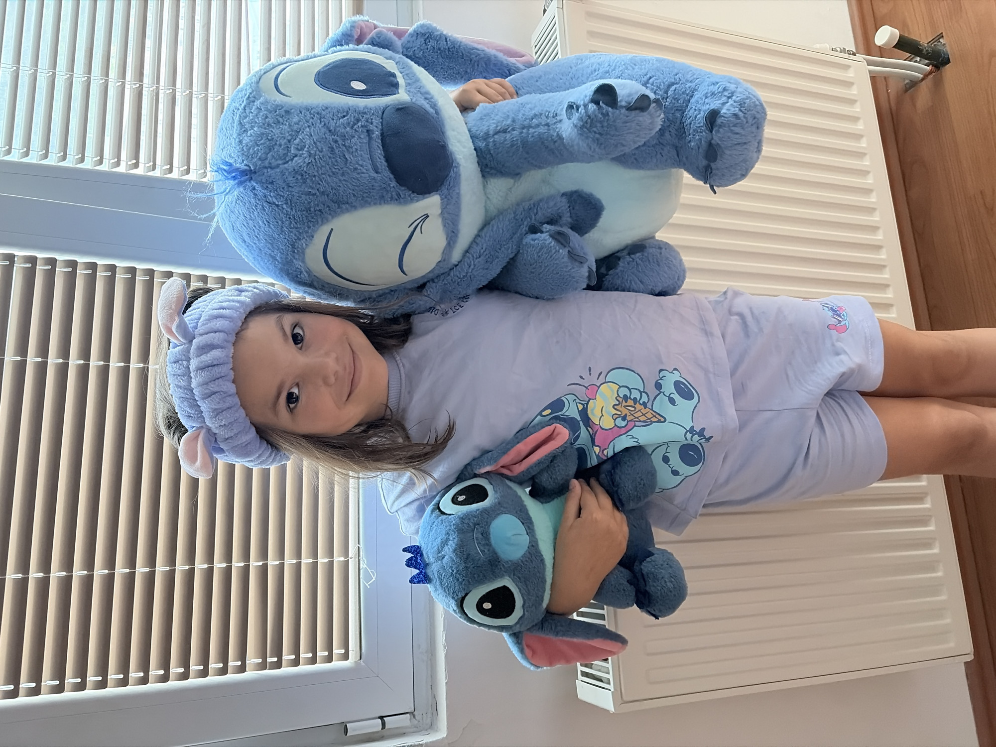 A young girl is lying on a white radiator cover, holding a smaller Stitch plush toy while a larger Stitch plush sits on top of her. She wears a lavender pajama set and a matching headband with bunny ears. The background includes window blinds and a partially visible wooden door. The location is likely a residence in the city of Budva, Montenegro.