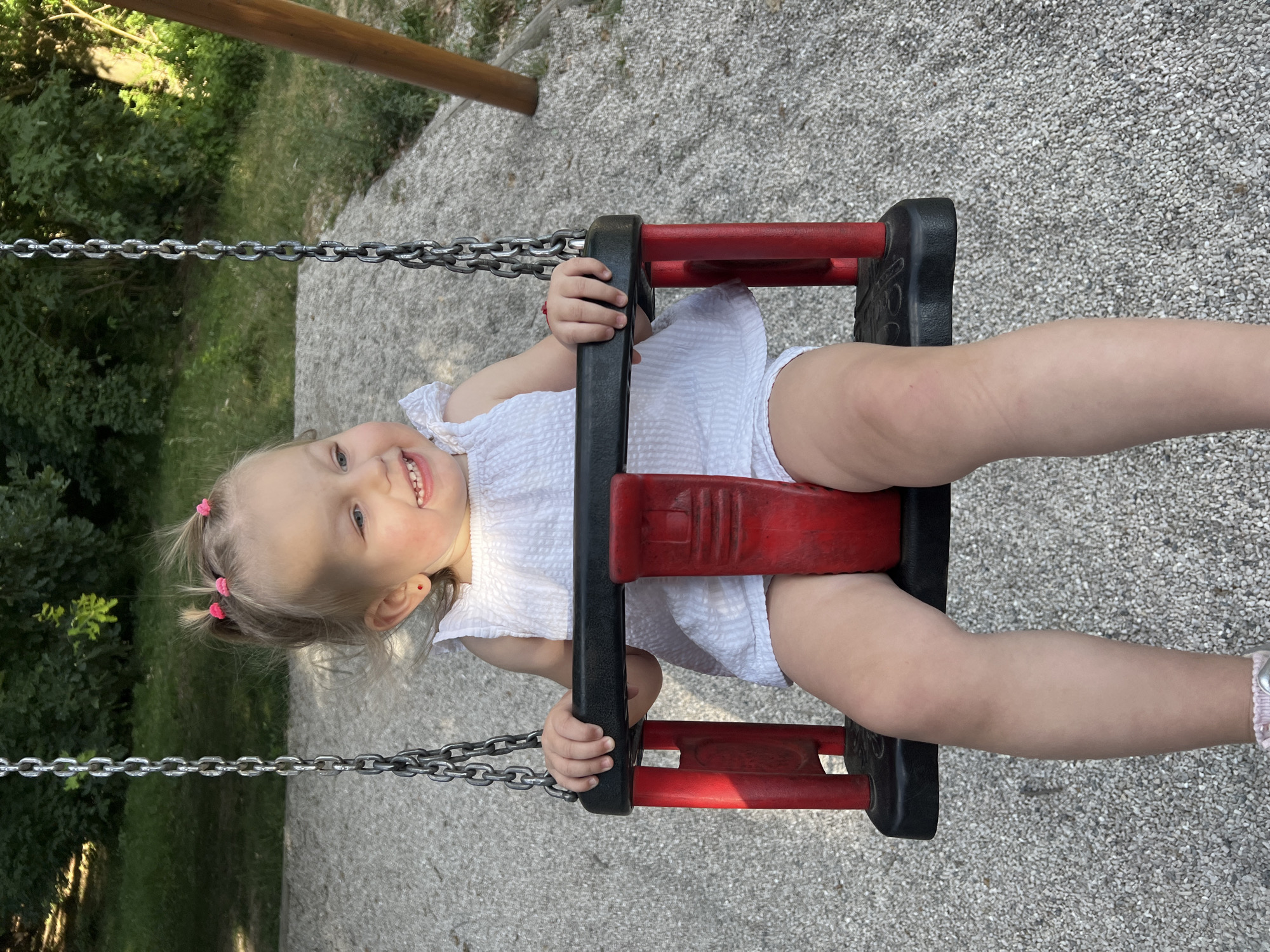 A young child with blonde hair in two pigtails is sitting on a red and black swing. She is wearing a white dress. The child is smiling and looking to the right. The swing is suspended by metal chains. The ground beneath the swing is covered in small, gray gravel. In the background, there are green trees and foliage, and a wooden pole is visible to the right. The scene appears to be in an outdoor playground.