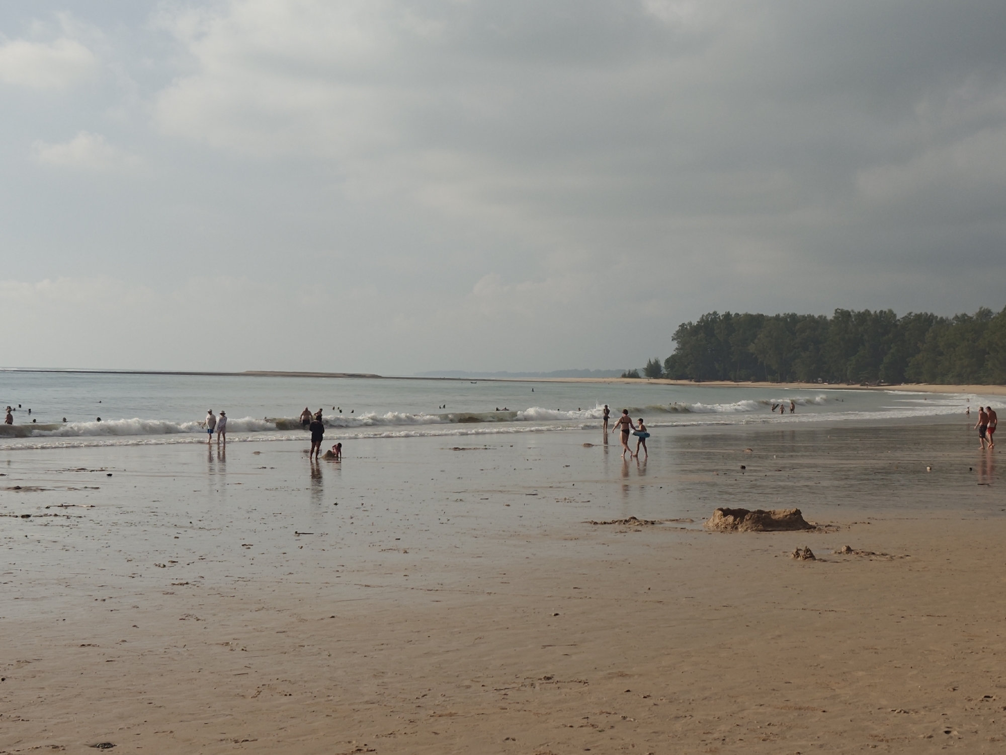 The image depicts a broad, sandy beach in Sakhu, Thailand, under an overcast sky. The ocean is calm with gentle waves breaking on the shore. Several people are visible in and near the water, some wading, others playing. On the sand, individuals are scattered, with a few appearing to be children playing. A small sandcastle or mound of sand is present in the foreground. In the distance, a line of trees forms the edge of the land. The overall impression is a relaxed beach scene on a moderately cloudy day.