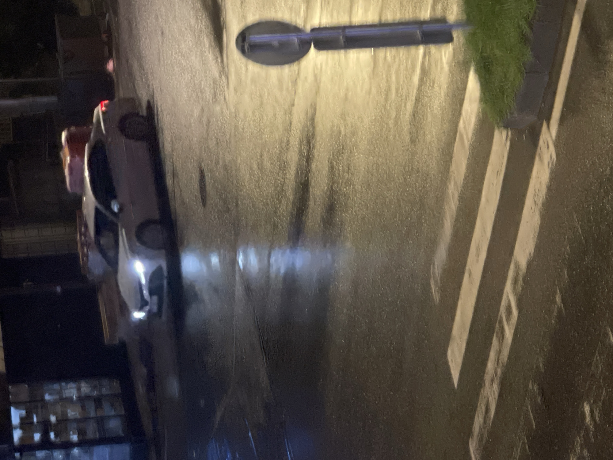 A car is driving at night on a wet street in Batumi, Georgia. The car's headlights are on. The street has a crosswalk. There is a traffic sign on the right side of the image, and a patch of grass next to it. The scene appears to be in an urban area, with buildings visible in the background. The overall lighting is dim, with reflections on the wet road surface.