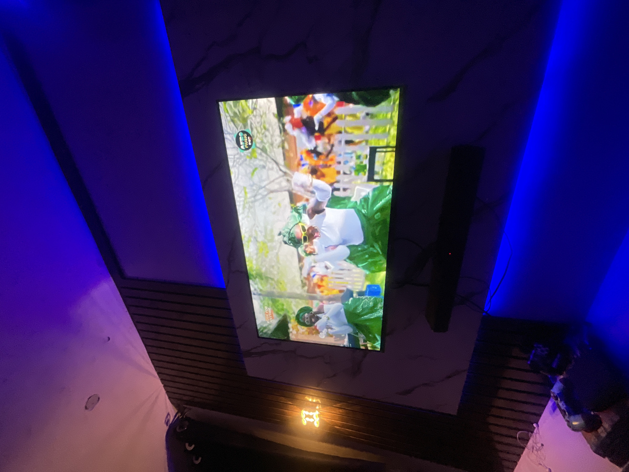 The image shows a wall-mounted television in a room with distinctive blue and purple lighting accents. The television displays a video featuring people in white clothing and green foliage, possibly a music video given the "MTV Music" logo in the upper left corner of the screen. Next to the television on the wall is a black, cylindrical speaker. The wall behind the television appears to have a marble-like finish. Below the television, there is a dim light fixture. In the foreground, parts of the room are visible, including the floor, a small table with various objects on it, and a white object on the floor. The image does not contain enough information to determine the location in Lokoja, Nigeria.