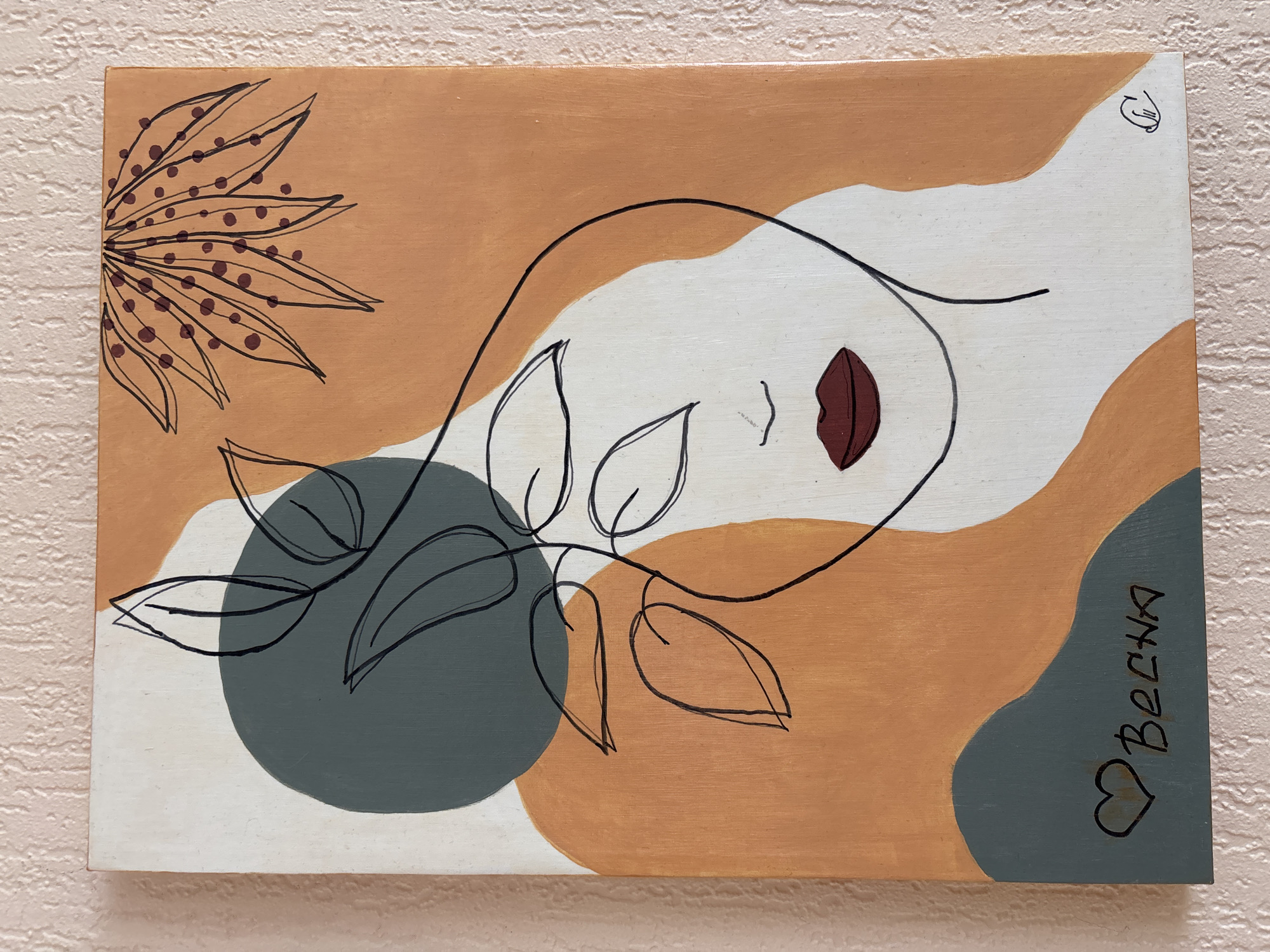 The image displays a painting on a canvas depicting a minimalist face with dark outlines and a few colored shapes. The face is drawn with a single continuous line, with closed eyes and red lips. Abstract shapes of muted orange, grey, and white fill the background. In the upper right corner, there is a stylized drawing of a leaf with dark dots. In the lower left corner, there is a heart symbol and the word "Весна" (Vesna, meaning Spring in Russian) written in script. A small artist's signature, a stylized 'G', is located in the lower right corner. The canvas is leaning against a textured, light pink wall. The painting is located in the city of Krasnodar, Russia.
