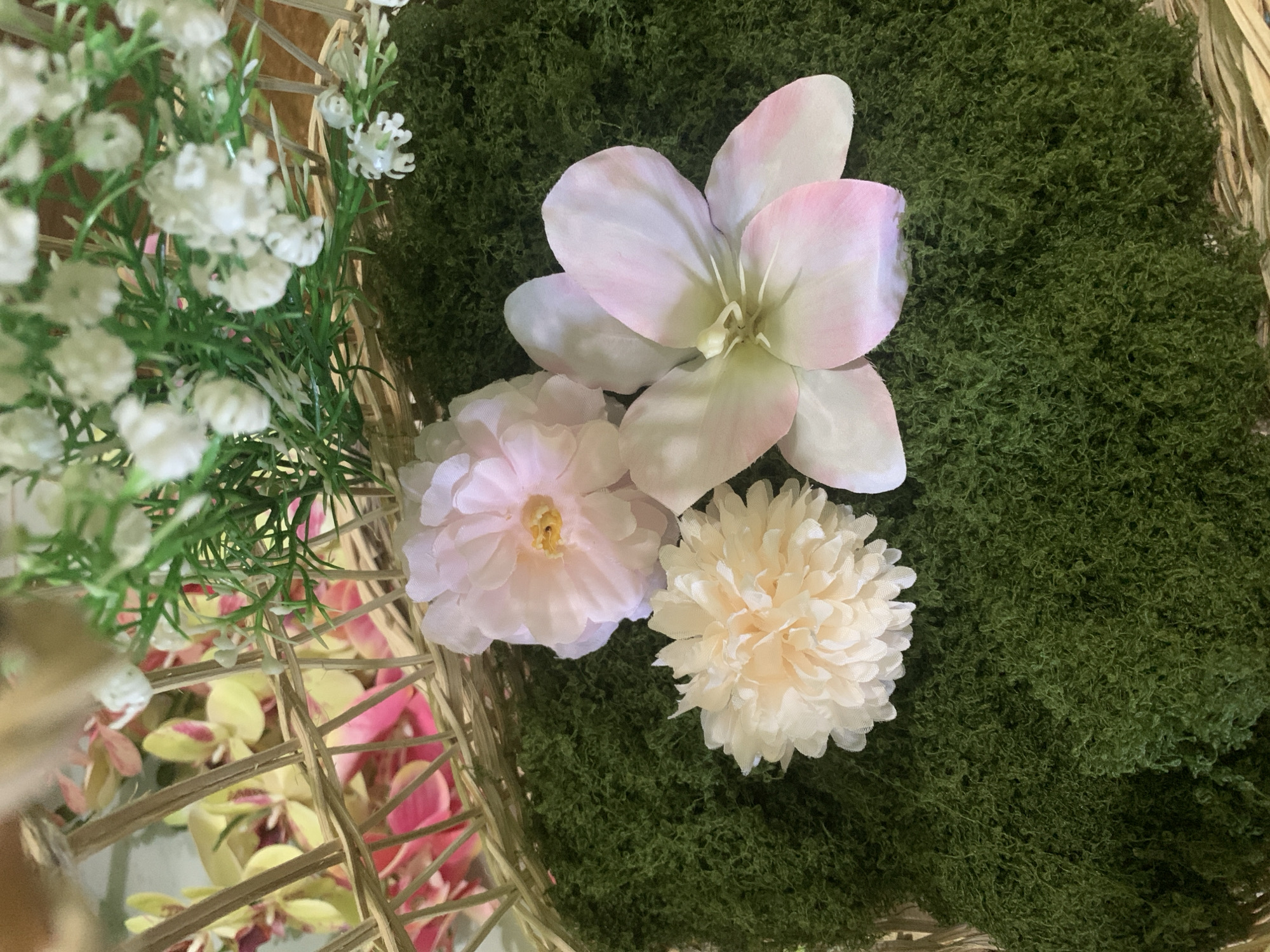 The image depicts a close-up view of artificial floral arrangements placed within a woven basket. The basket's natural, light brown material forms the background and frame for the foreground elements. A thick layer of dark green, textured moss covers a portion of the basket's interior. Resting on the moss are three artificial flowers. The largest flower, positioned on the right, is light pink with white accents and has five prominent petals. To its left and slightly above is a pink flower with multiple layers of soft, ruffled petals and a yellow center. Below and to the left of the pink flower is a cream-colored, spherical flower composed of numerous fine, delicate petals. In the upper portion of the image, behind the moss and flowers, are additional artificial plants. These include a cluster of small, white, star-shaped flowers mixed with feathery green foliage, and further back, a section of a plant with pointed yellow and pink leaves woven into the basket. The lighting is soft, casting gentle shadows on the moss and flowers, highlighting their textures and colors. There are no people or animals present in the scene. The image is a still-life arrangement of decorative artificial plants and flowers.