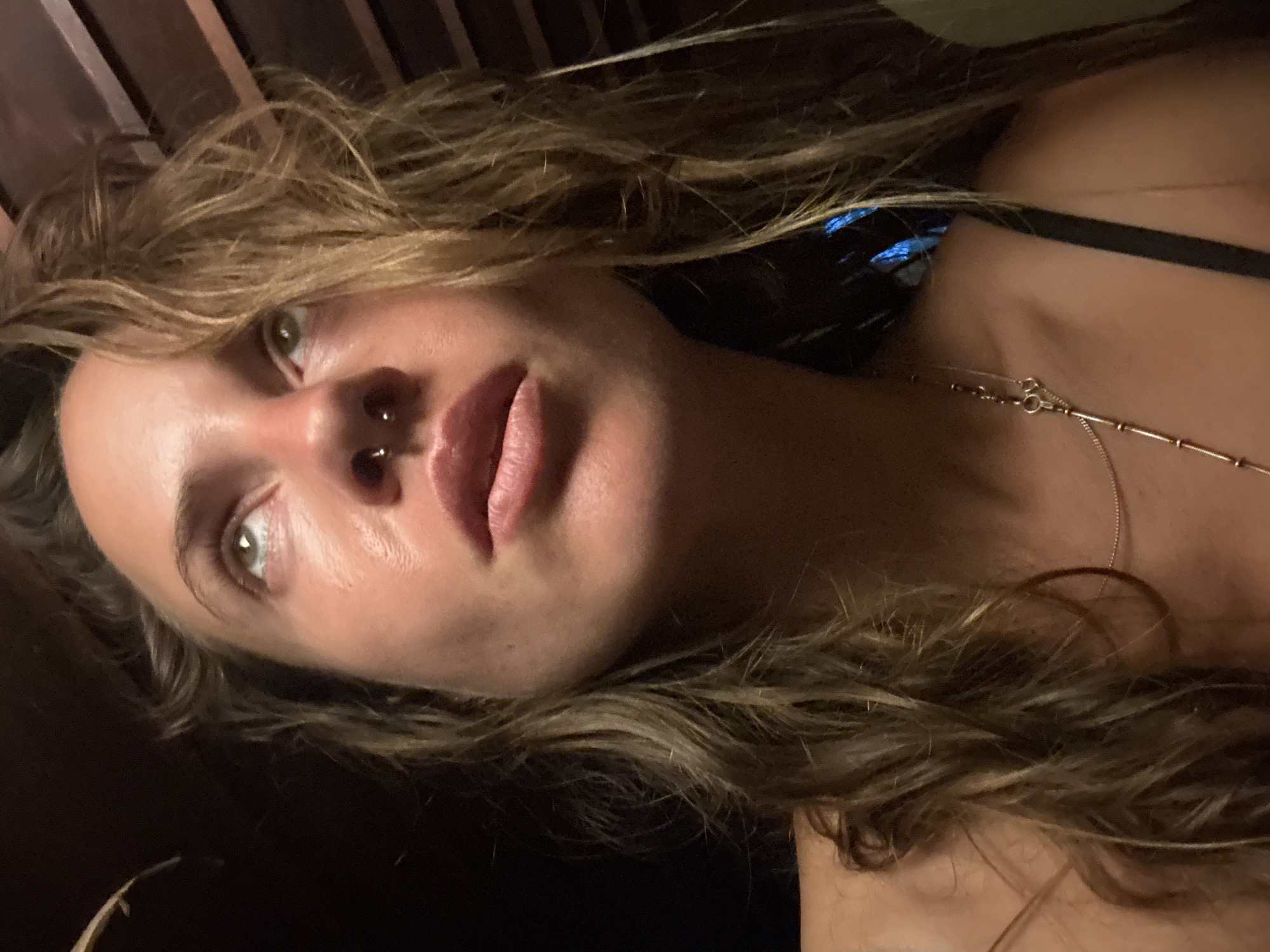 A close-up, low-angle, indoor shot features a young adult with light brown, wavy hair looking upwards with a slightly parted mouth. The subject has a septum piercing. Their skin appears sun-kissed and slightly shiny. They are wearing a thin strap that is part of an undergarment or dress. A delicate silver necklace with small bead-like details is visible on their décolletage. The background is dark with hints of wooden beams overhead, suggesting an interior space with some architectural detail, potentially a covered patio or room with a slatted ceiling. The lighting is dim and appears to be coming from above and slightly to the side, casting subtle shadows on the subject's face.