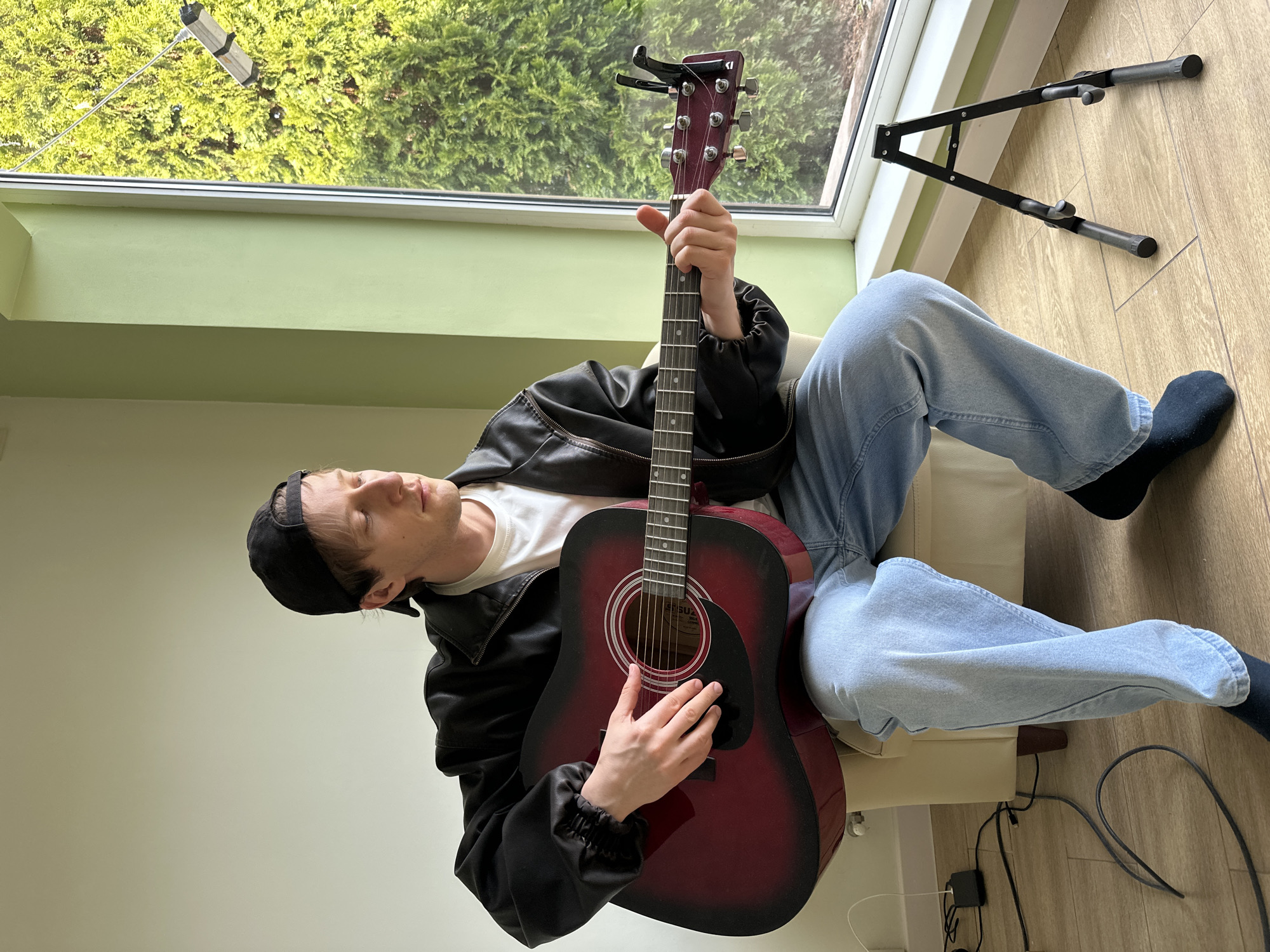 The image depicts a young man seated indoors, holding a red acoustic guitar. He wears a black baseball cap, a white t-shirt under a black jacket, and light blue jeans. His right hand is positioned on the guitar's sound hole, while his left hand grips the guitar's neck. He appears relaxed with his eyes closed.
Behind him, a large window reveals lush green foliage, likely trees. To the right of the window, a black guitar stand is visible on the floor. Miscellaneous cables and a power adapter are also on the floor near the chair.
The location is not possible to determine to be the city of Kursk, Russia from the image alone. The environment suggests an interior setting with natural lighting from the window.