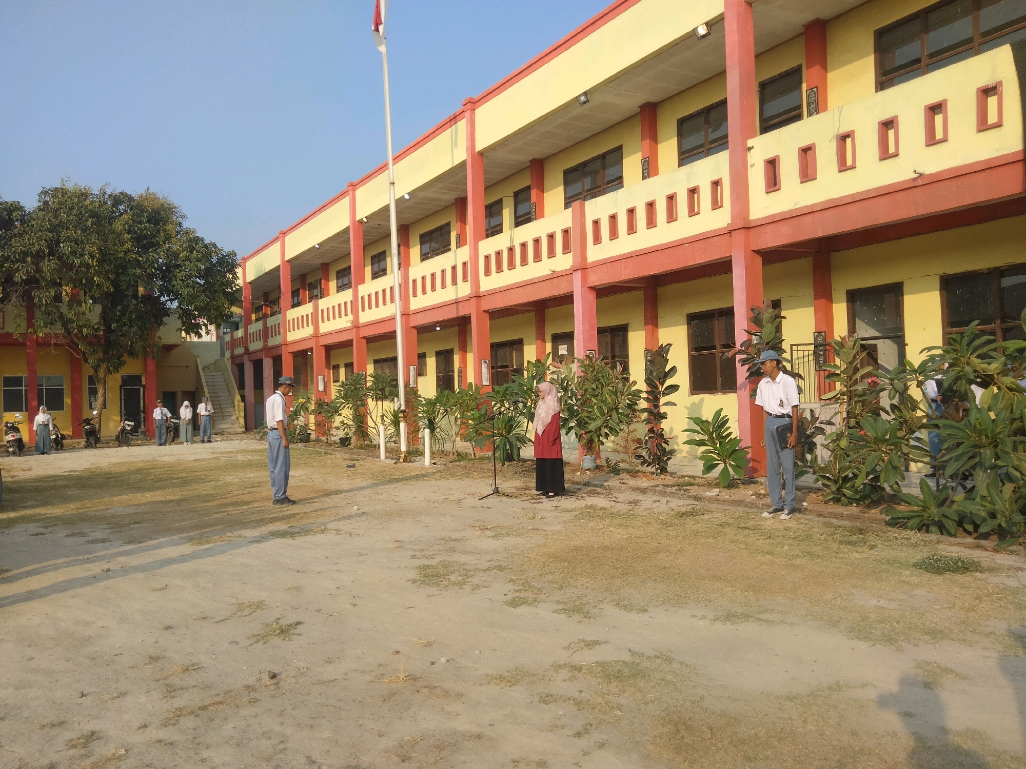 The image captures an outdoor scene in front of a yellow and red two-story school building in Suka Sari, Indonesia. The building has a red-painted facade with yellow walls. Several students in school uniforms are present. On the right, a male student wearing a white shirt, blue pants, and a gray cap stands holding a black folder. In the center, a female student in a pink hijab and a red top over a dark skirt stands in front of a microphone. To the left, another male student in a white shirt and blue pants stands near the building entrance. Further left, a group of students, some wearing hijabs, are gathered near motorcycles parked in front of the building. The ground is covered with dirt and patches of dry grass, and shadows are cast by the building and trees. A flagpole with an Indonesian flag is visible in the background. The overall atmosphere appears to be during an outdoor school event or ceremony.