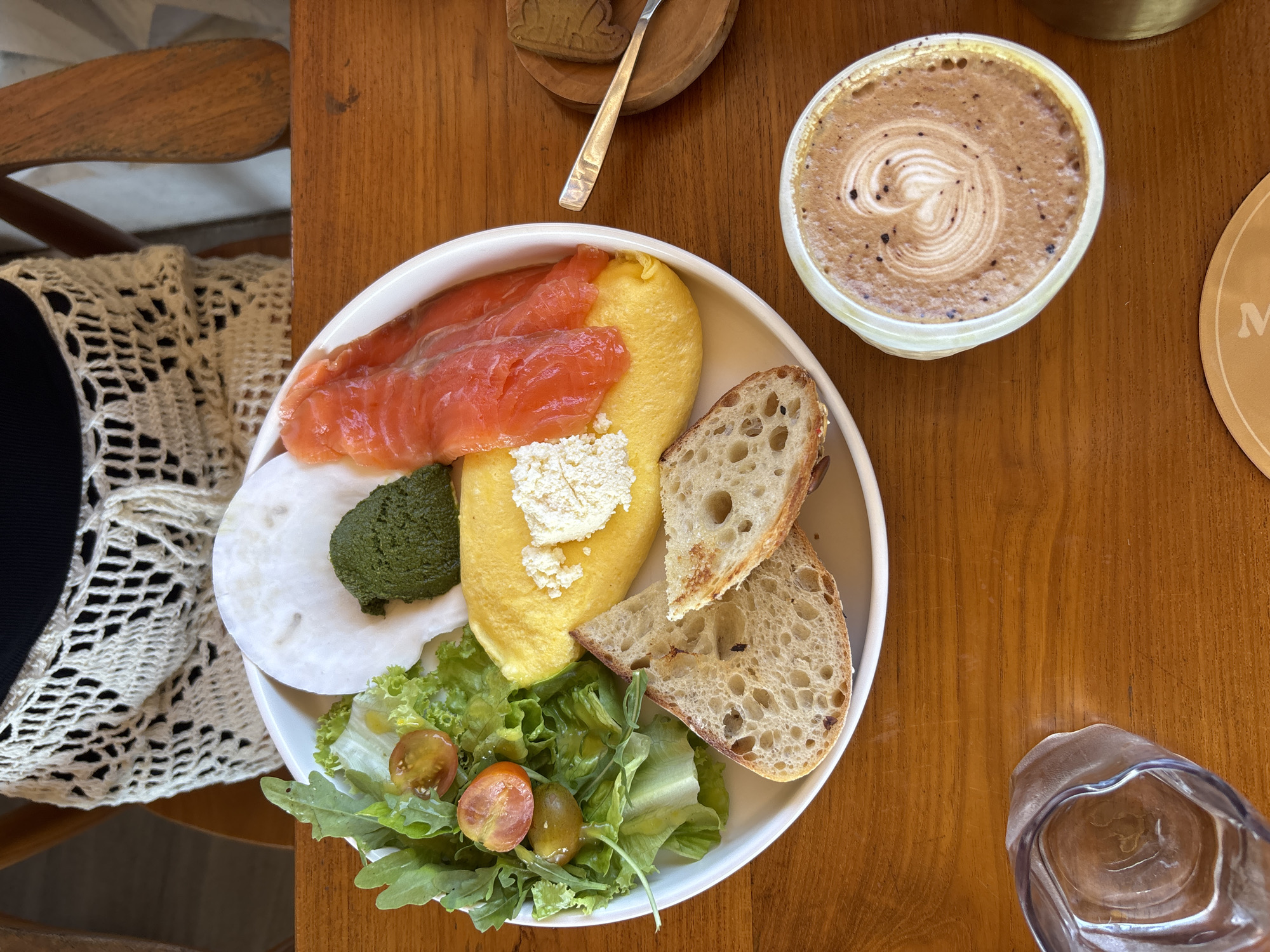 A high-angle shot shows a breakfast meal on a wooden table, possibly inside a café or restaurant. The meal includes smoked salmon, eggs with white cheese crumbles, a green spread, a white vegetable slice, salad with halved cherry tomatoes, and two slices of sourdough bread arranged on a white plate. Beside the plate, there is a cup of cappuccino with latte art. A water glass is on the bottom right. The presence of an intricate crochet sweater and a wooden chair suggests a person is seated at the table. The location is unspecified within Kuta Selatan, Indonesia.