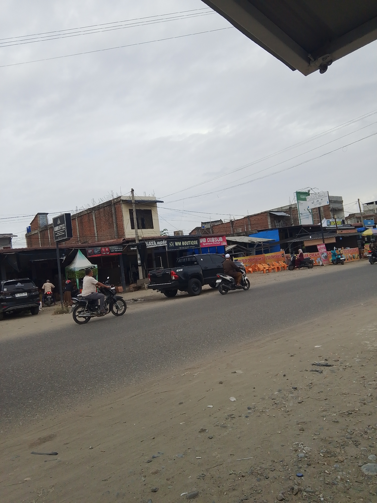 This is a photo of a street scene in Gampongpaya, Indonesia. The time of day appears to be late afternoon or early evening, given the overcast sky and muted lighting. The weather is cloudy, contributing to the subdued atmosphere.
The main subjects are several people on motorcycles, driving on a paved road. There are also parked cars, including a black SUV with a license plate that reads "BL 1692 KH." In the background, there is a row of shops and buildings lining the street. Some of the shops have awnings and signs, indicating various businesses. Visible text on the signs includes "AMRI STYLE," "KAYA mode," "WM BOUTIQUE WM DIMSUM," and "OPPO." There are also signs advertising food items like "STELER CREAM DURIAN." Orange plastic chairs are set up outside some of the establishments, suggesting a place for customers to sit. The overall scene depicts a typical commercial street in what appears to be a moderately busy area. The ground in the foreground is unpaved and sandy, with scattered debris.
