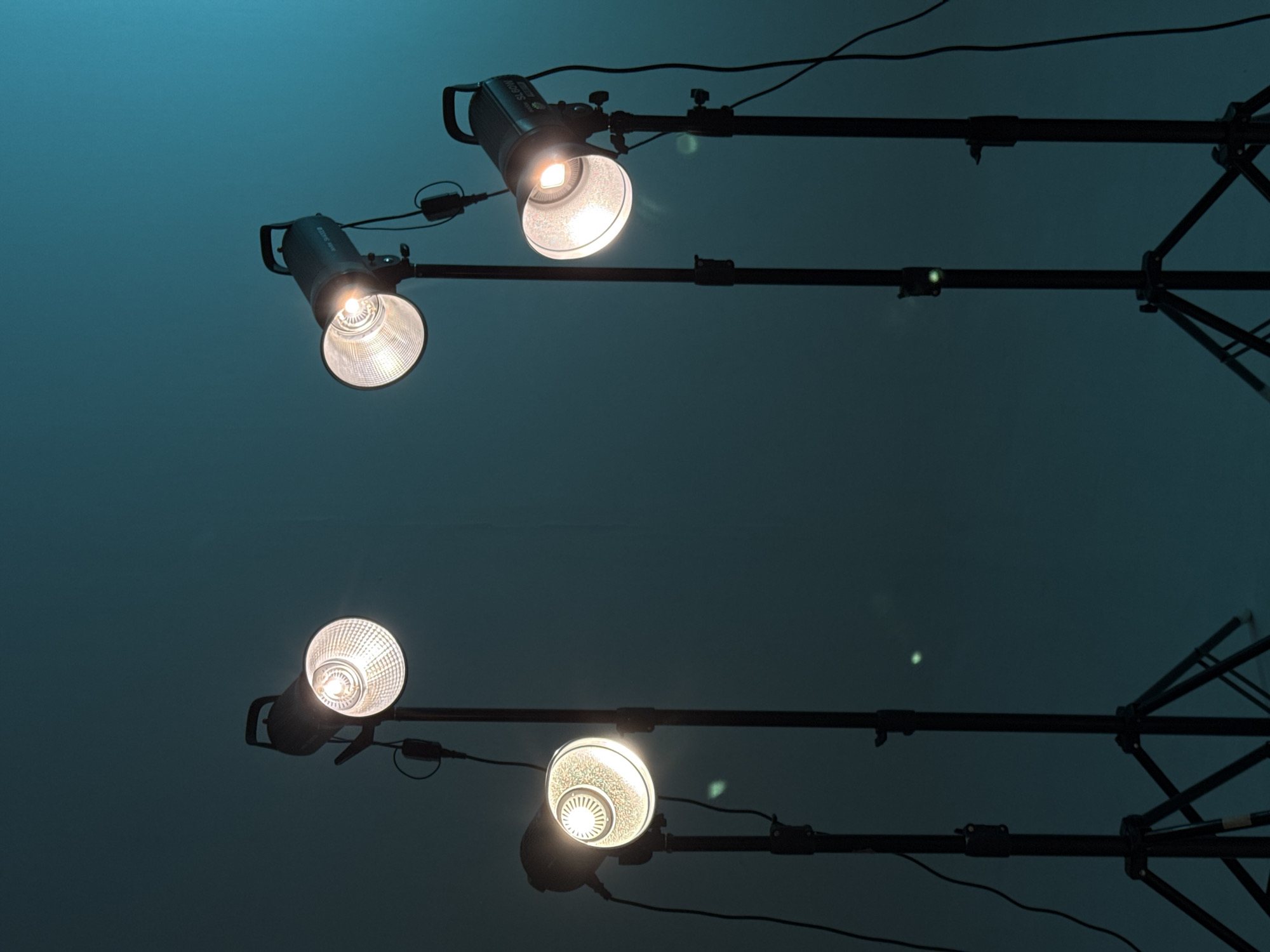 The image depicts four studio lights attached to stands, illuminating a dark teal or blue-green background. The lights are of varying designs, some with reflective interiors and others with textured diffusers. They are positioned at different heights and angles. Black power cables and supports are visible against the colored backdrop. The photograph does not provide information to infer location.