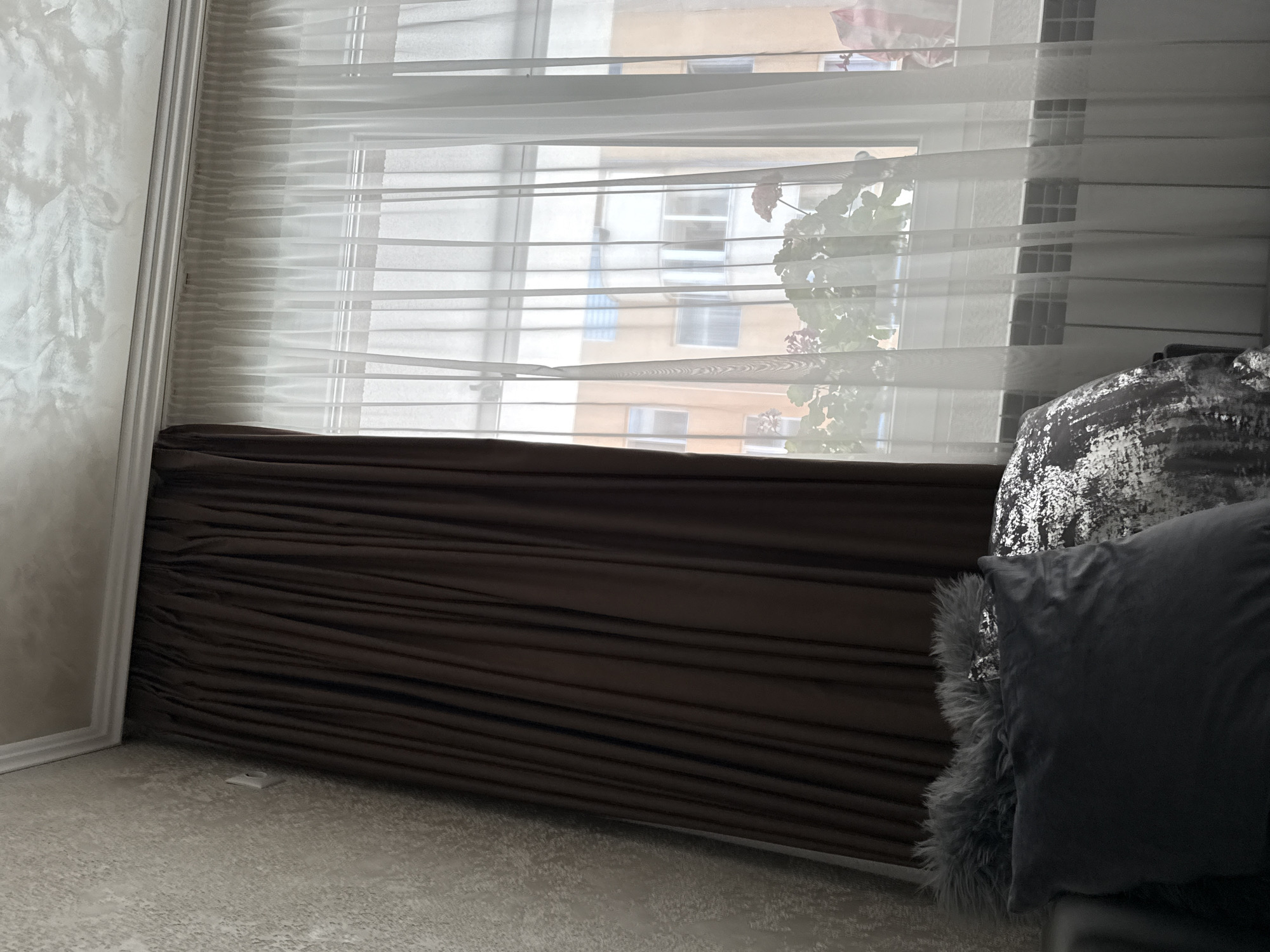 The image depicts an interior scene focused on a window with curtains and pillows in the foreground. On the left, a dark brown curtain is drawn closed, obscuring a portion of the window. To its right, a sheer white curtain is drawn back, revealing the window and the exterior. Through the sheer curtain, a building with multiple windows and balconies is visible, suggesting an urban setting. A radiator is positioned below the window. In the foreground, on a dark surface, there are several pillows: a large dark grey pillow, a pillow with a shimmery, textured pattern, and two smaller grey furry pillows stacked behind them. The wall on the left has a textured finish, and the ceiling has a decorative molding and a textured finish. No people are visible in the image. The scene appears to be a residential interior, likely in Veliko Tarnovo, Bulgaria, given the architectural style of the building visible through the window.