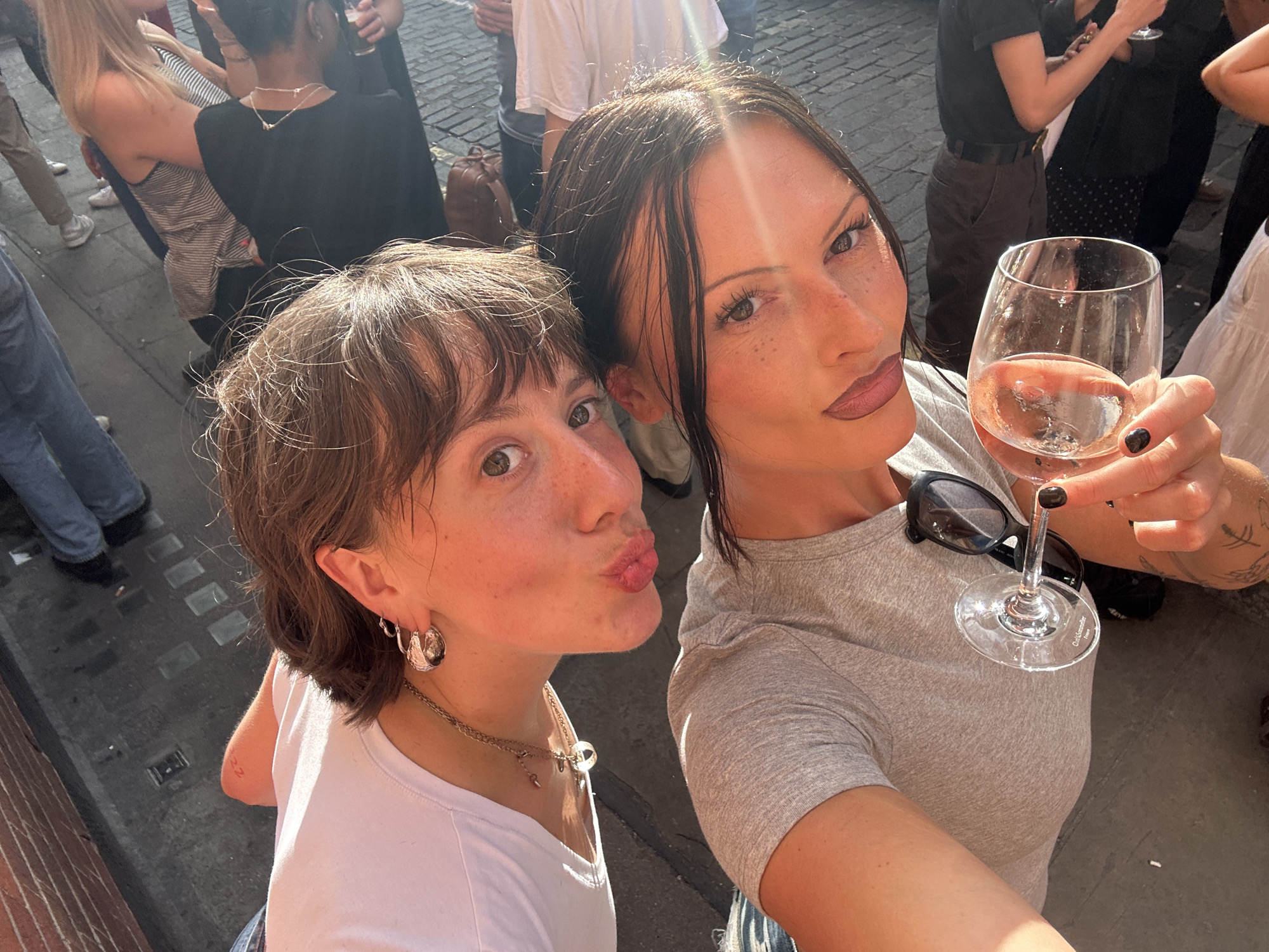 Two young women are posing for a selfie outdoors on a street in London. The woman on the left has short brown hair and is wearing a white t-shirt and hoop earrings. The woman on the right has long dark hair and is wearing a grey t-shirt. She is holding a glass of rosé wine and has sunglasses perched on her shirt. The background shows a cobblestone street and other people enjoying the sunny day. The scene suggests a casual social gathering in a bustling urban environment.