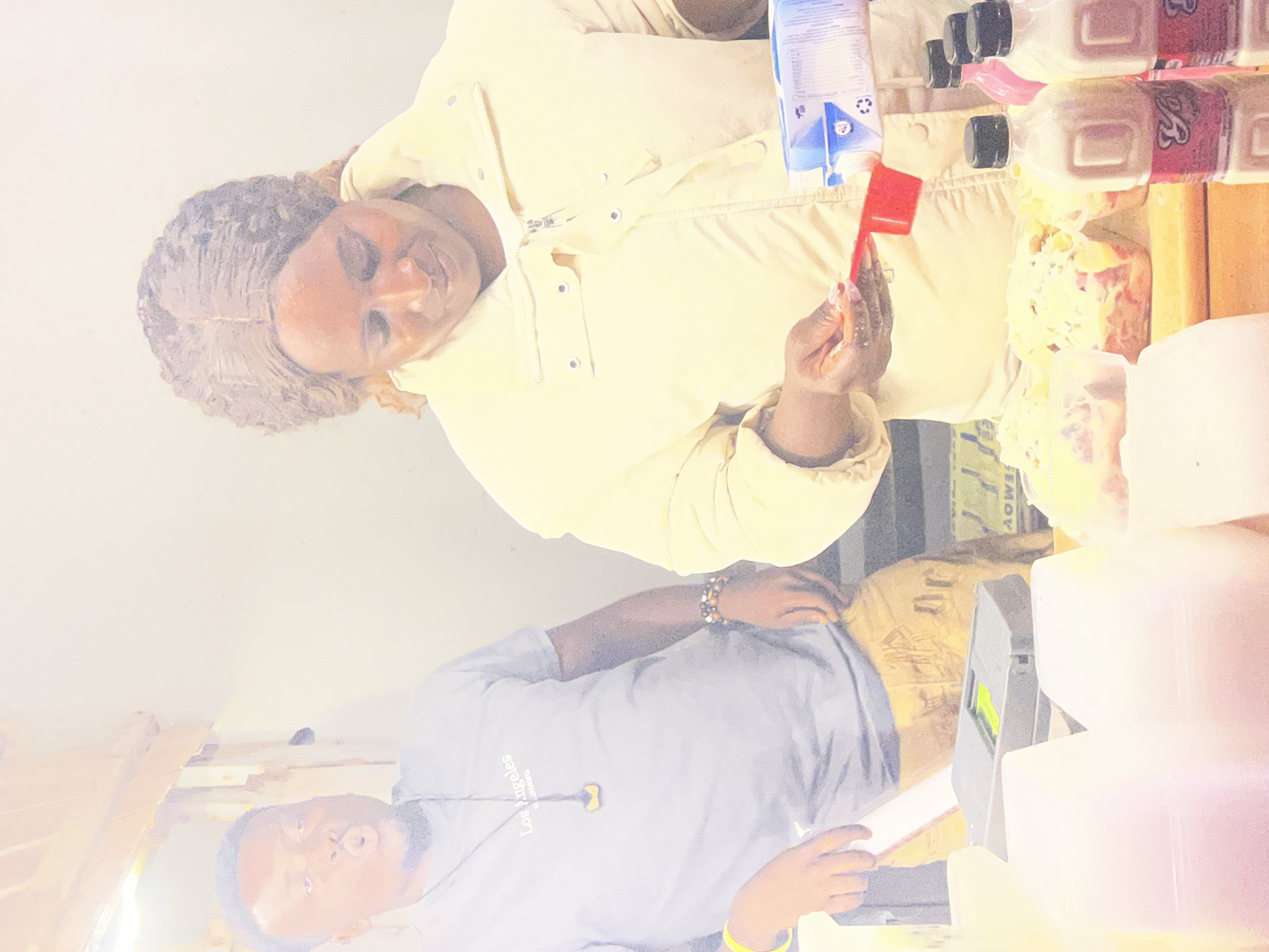 The image depicts an indoor scene, likely a food stall or a kitchen, with two individuals present. On the left, a man wearing a light blue t-shirt with "Los Angeles" printed on it and a pendant around his neck is captured with his mouth open in an exaggerated expression. He has a beard and is wearing a yellow wristband on his left arm. To his right, a woman wearing a light yellow puffy jacket is focused on an action. She is holding a red measuring scoop in her right hand, pouring a white liquid from a blue and white carton into it. The carton appears to be for milk or a similar beverage. In the foreground, there are several white containers, a digital scale with a green section, and some food items, possibly pastries or desserts, arranged on a wooden surface. Several plastic bottles with black caps and pink and white labels are also visible. The overall lighting suggests an indoor environment with artificial light sources. The image does not provide any specific visual cues to pinpoint the location as Durimi, Nigeria.