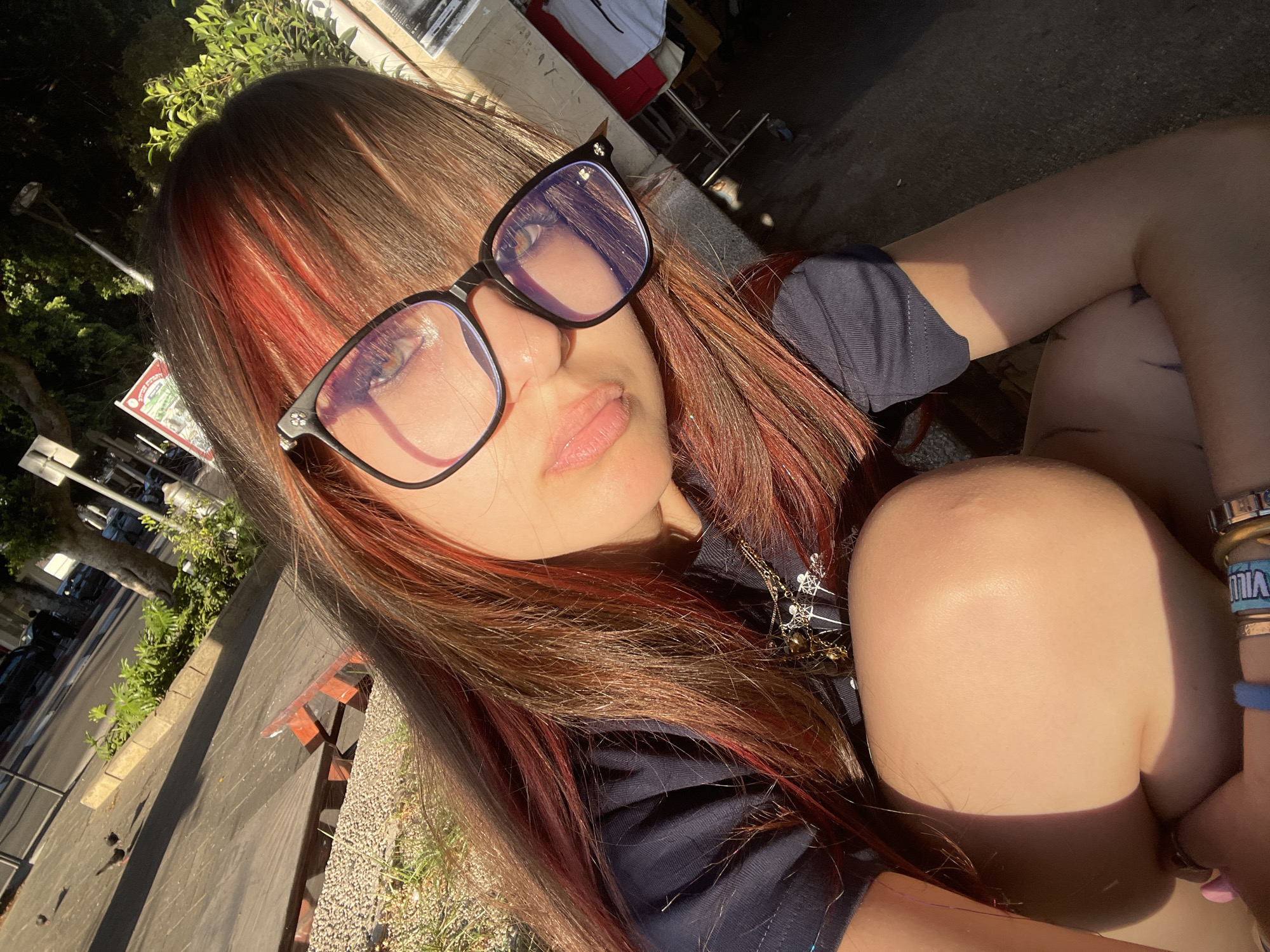 A young woman with reddish-brown hair and glasses is taking a selfie outdoors in Raanana, Israel. She is wearing a dark blue t-shirt and has her knee close to her chest. Sunlight casts shadows on her skin. In the background, there are trees, a sidewalk, and a street. Some buildings are also visible.