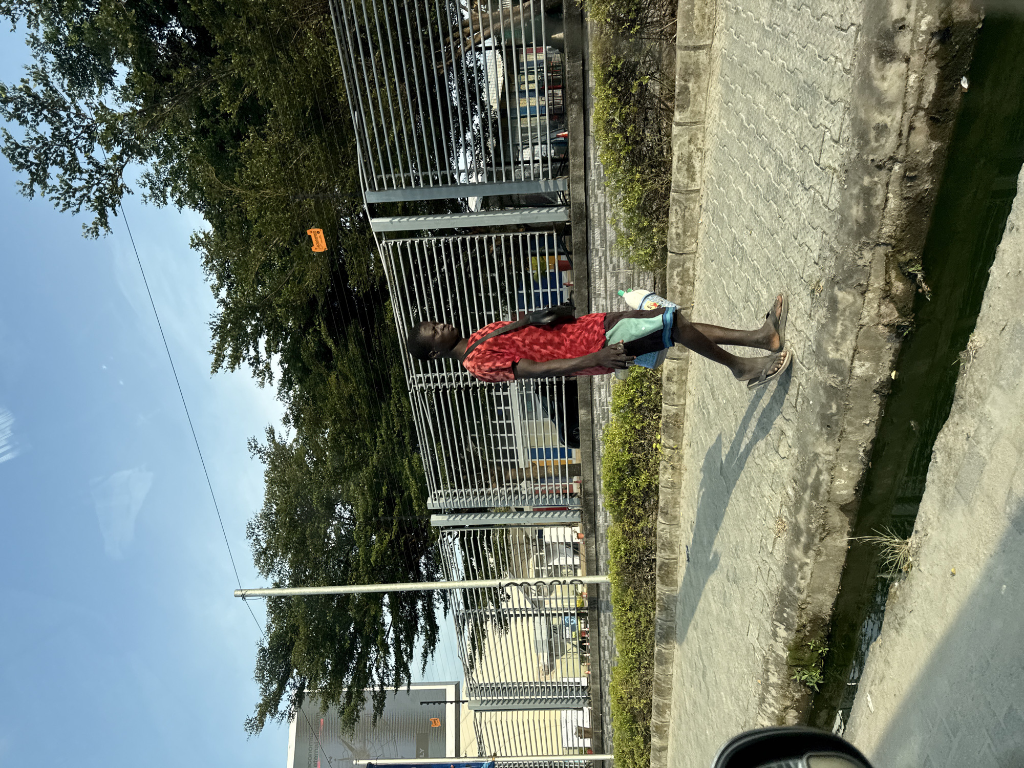 The image shows a young Black man walking along a paved sidewalk next to a drainage ditch. He is wearing a red t-shirt, patterned shorts, and flip-flops. A black bag is slung across his chest, and he is carrying some light-colored items in his left hand. Behind him is a metal fence with horizontal bars, and beyond that, trees and a building with a large billboard. The sky is clear and blue. The scene is consistent with an urban or semi-urban environment in Igboefon, Nigeria. The lighting suggests it is daytime, likely late afternoon given the long shadow cast by the man.