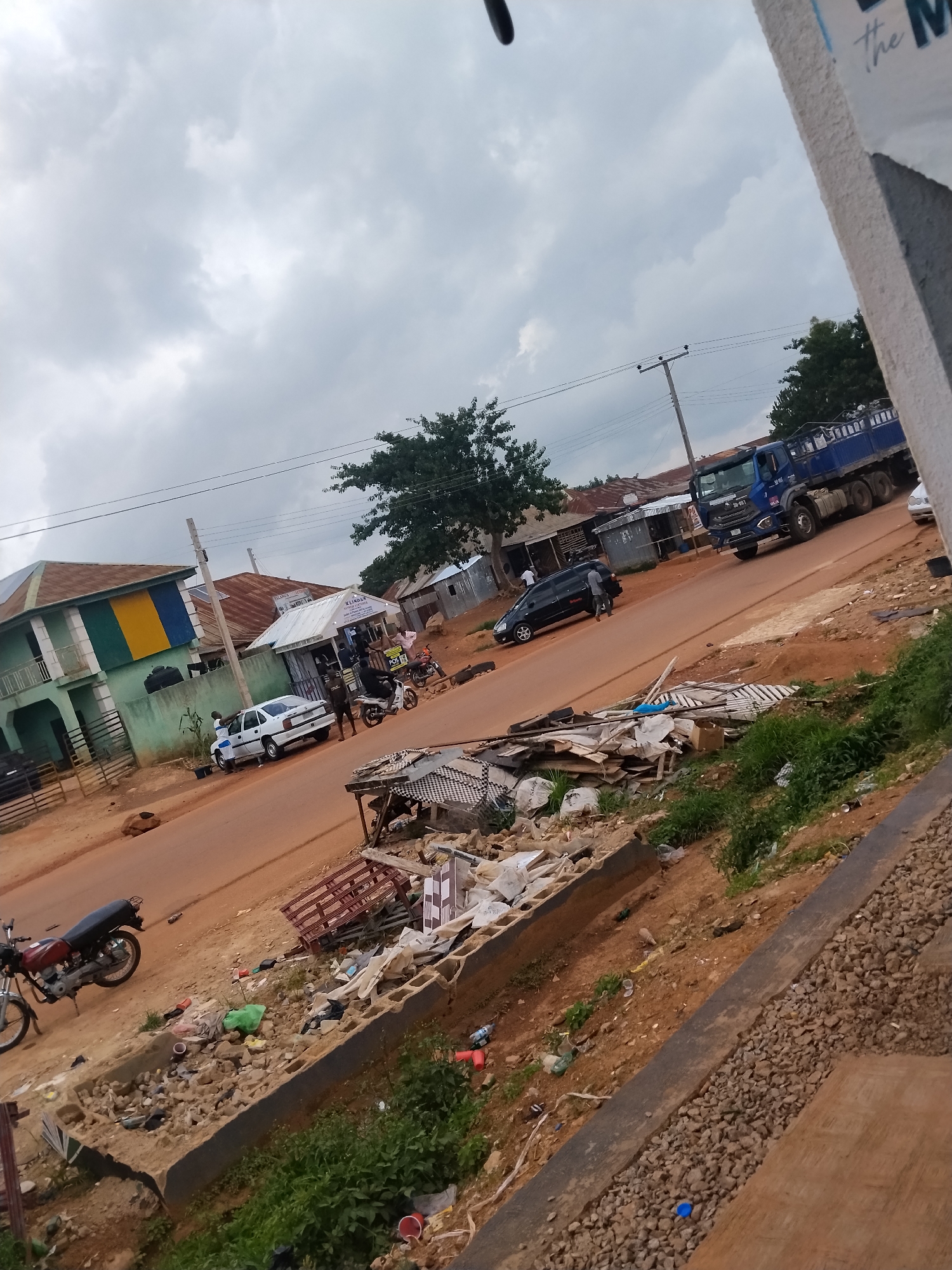 This image captures a street scene in Durumi, Nigeria, under a cloudy sky, suggesting it could be late afternoon or an overcast day. The foreground shows a pile of debris, including broken concrete blocks, plastic waste, and some metal. A red and black motorcycle is parked on the side of the dusty, reddish-brown road.
Further down the road, a white car is parked in front of a building with a green and yellow facade. Next to it, a small commercial building with a white awning has several people standing outside and a motorcycle parked nearby. Across the street, a large blue truck is driving away from the camera. Black cars are also visible on the road.
The overall setting appears to be a moderately busy commercial and residential area. The presence of various vehicles and people suggests daily life and economic activity. The debris in the foreground might indicate ongoing construction or waste disposal issues in the area. There is no discernible text in the image that provides specific information about businesses or locations.