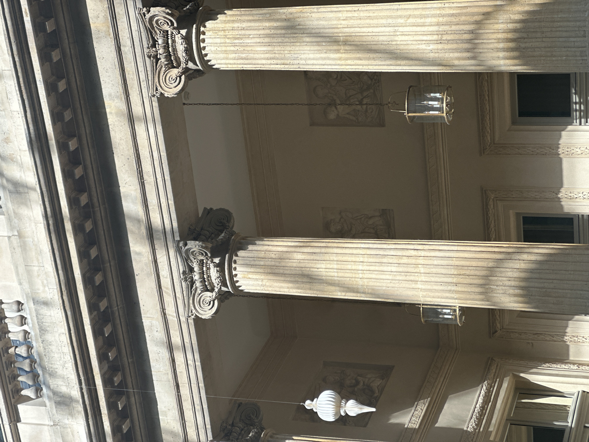 The image shows the ornate architectural details of a building exterior, likely located in Paris, France. The focus is on the classical elements: fluted columns with Corinthian capitals, decorative friezes, and a series of windows recessed within the building's façade. Hanging lights, some cylindrical with visible bulbs, are suspended between the columns. Light and shadow play across the textured surfaces, highlighting the intricate carvings and molding. The overall impression is one of grand architectural design and historical significance.