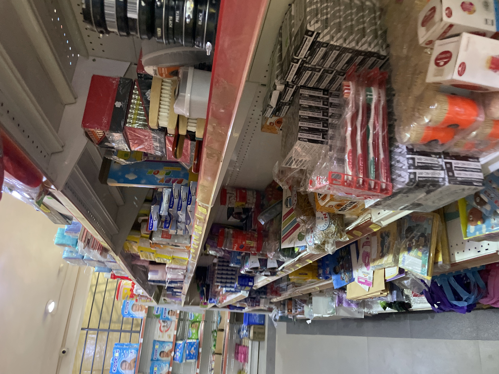 The image depicts shelves in a retail store located in Kurunduma, Nigeria. The shelves are stocked with a variety of merchandise, including shoe polish, brushes, toothbrushes, notebooks, baby products, and other packaged goods. The shelves are overloaded with merchandise, extending to the ceiling. The store is located indoors and lit with overhead lighting. There are no people visible in the image. The overall impression is that of a small, densely-packed retail outlet.