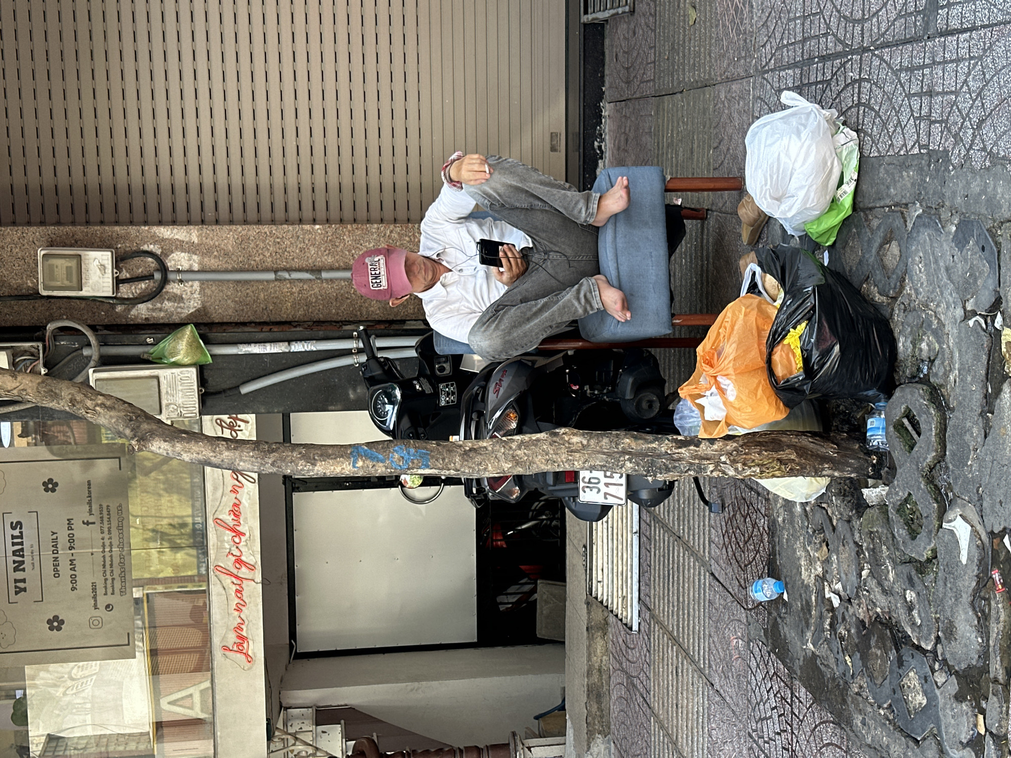 A man in a pink baseball cap and a white shirt sits barefoot on a blue cushion atop a parked black scooter in front of the "YI NAILS" salon in Thủ Đức, Vietnam. He is using a phone. The scooter is parked adjacent to the curb and close to a closed metal shutter door. Several bags of trash are visible on the sidewalk.