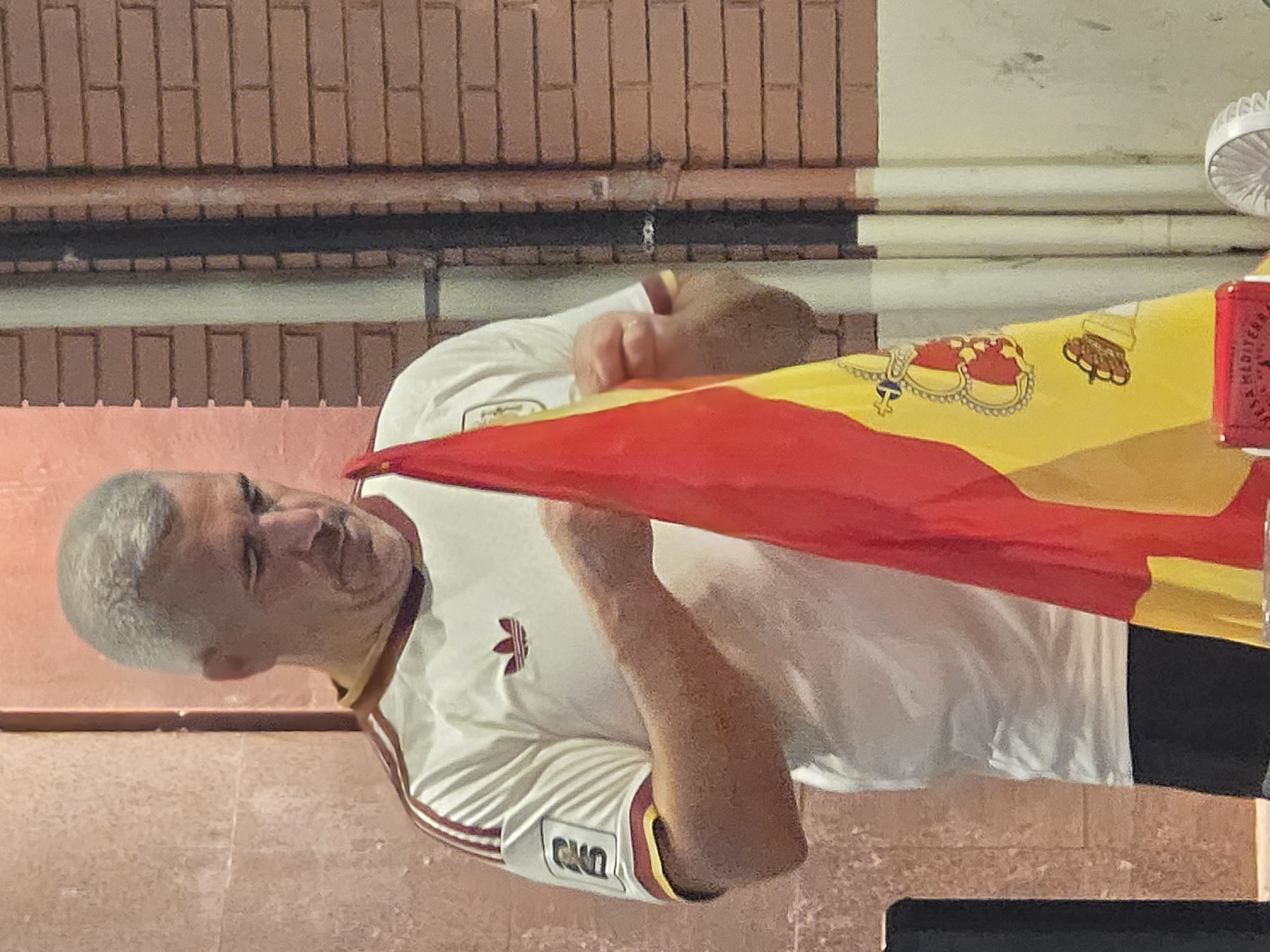 The image shows a middle-aged Caucasian man with graying hair and a mustache, wearing a white Adidas t-shirt with maroon stripes on the sleeves and a maroon collar, and dark shorts. He is holding a Spanish flag, which is predominantly red and yellow, with the coat of arms visible on the yellow portion. The man appears to be standing outdoors, with a textured pink wall behind him on the left and a brick wall with pipes on the right. The background suggests an urban or semi-urban setting. The lighting is somewhat dim, with a slight reddish cast. The overall scene appears to be casual and possibly related to a sporting event or a cultural gathering.