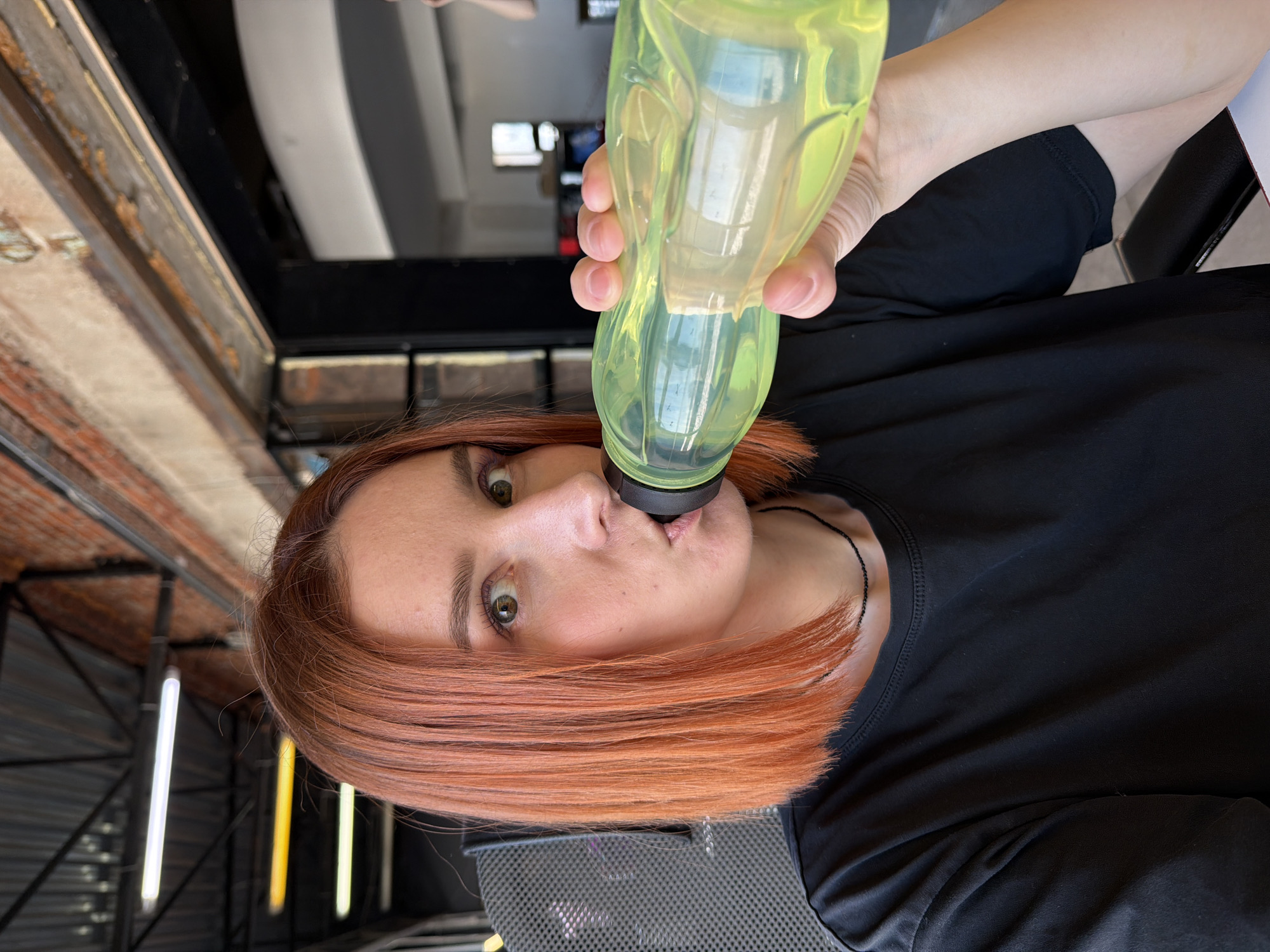 A woman with reddish-orange, shoulder-length hair is drinking from a translucent green water bottle. She is wearing a black t-shirt and appears to be indoors. In the background, elements of the interior architecture are visible, including what looks like metal framing and fluorescent tube lighting. The location appears to be in a building in the city of Chaikovsky, Russia.