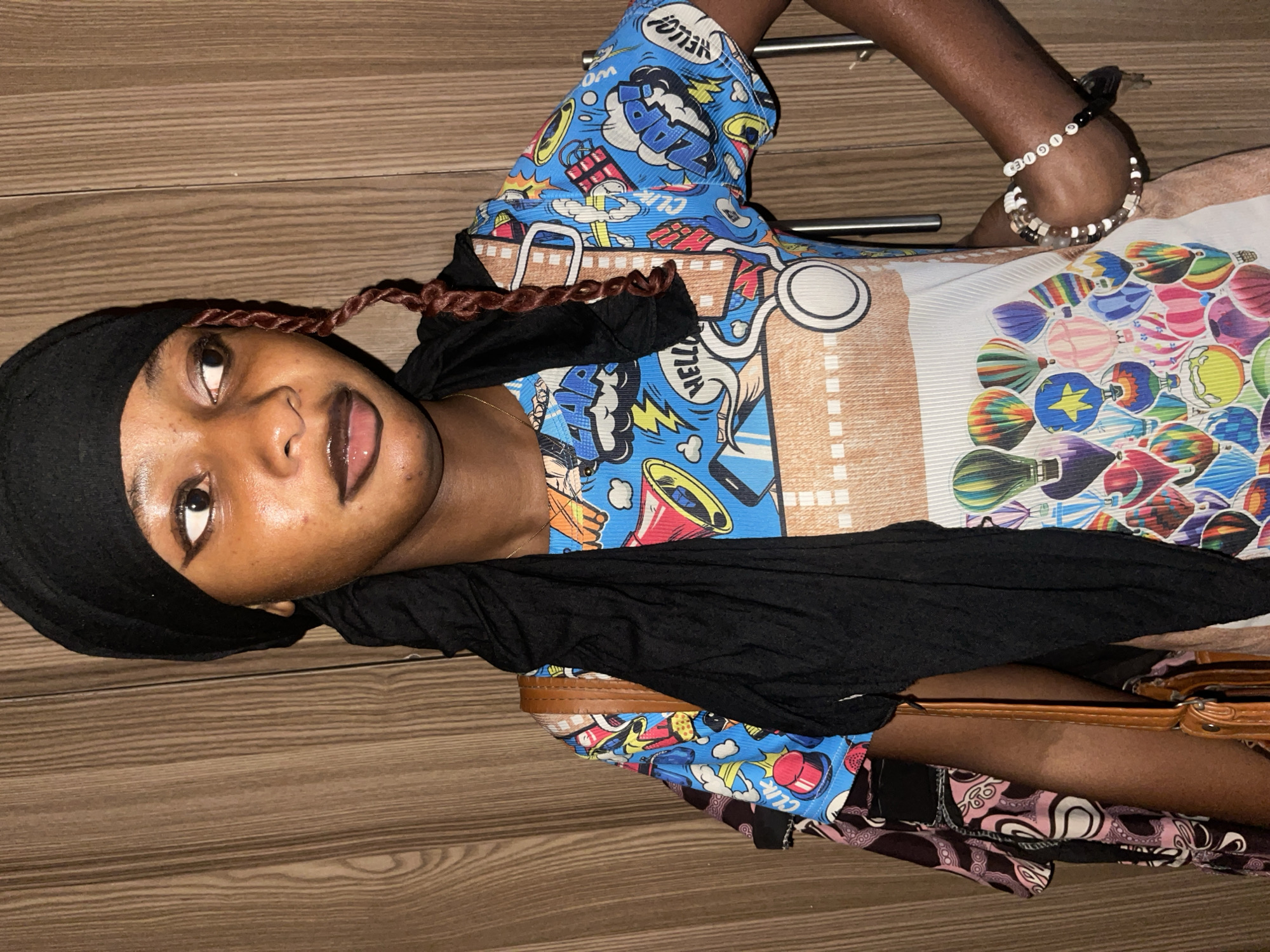 A medium shot depicts a dark-skinned woman wearing a black headscarf and a colorful, patterned shirt. She is lying on a light brown wood-grain surface. A black scarf is draped around her neck. Bracelets are visible on her left wrist. Part of a brown bag is visible near her lower body. The location is not discernible from the visual information provided.