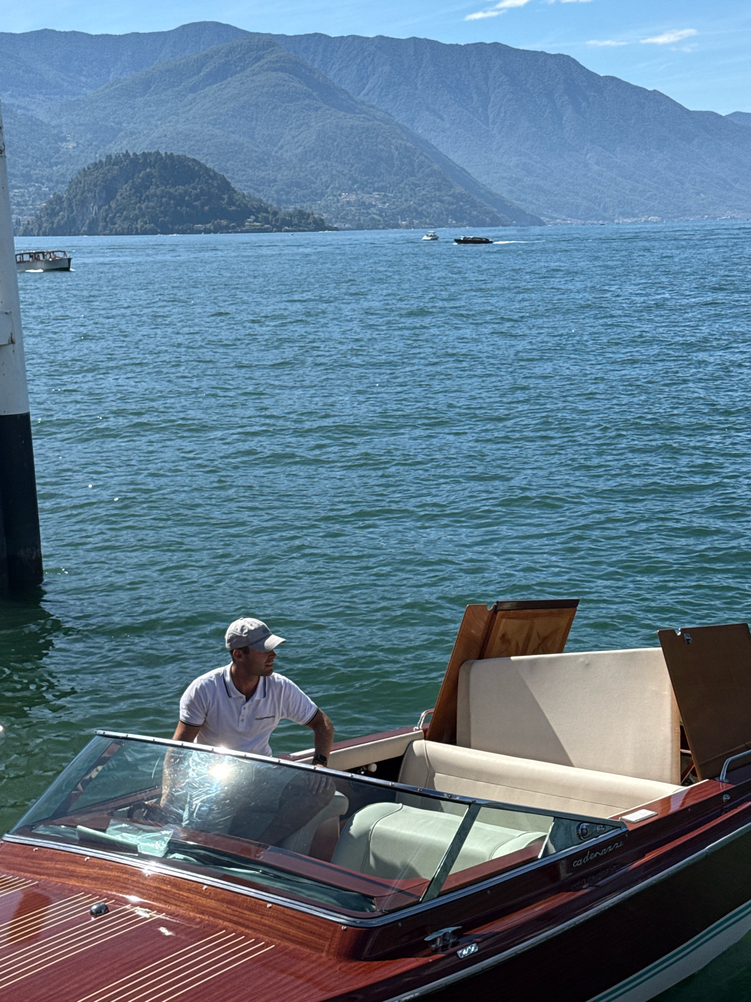This is a vertical, medium shot of a man standing on a wooden boat in Lake Como, near Varenna, Italy. The man is wearing a white polo shirt and a grey baseball cap, and he is looking to his right. He is standing next to the boat's windshield, which is reflecting the sunlight. The boat is a classic wooden runabout with light-colored seating. In the background, there are green hills and mountains under a clear blue sky with some white clouds. Several other boats are visible on the lake in the distance, including a small white one on the left and a darker, faster-moving boat further out. The water is a deep blue with small waves. A white and black post is visible on the left side of the frame, partially obscuring the view of the boat and the lake. The overall scene conveys a sense of leisure and beautiful scenery on a sunny day.
