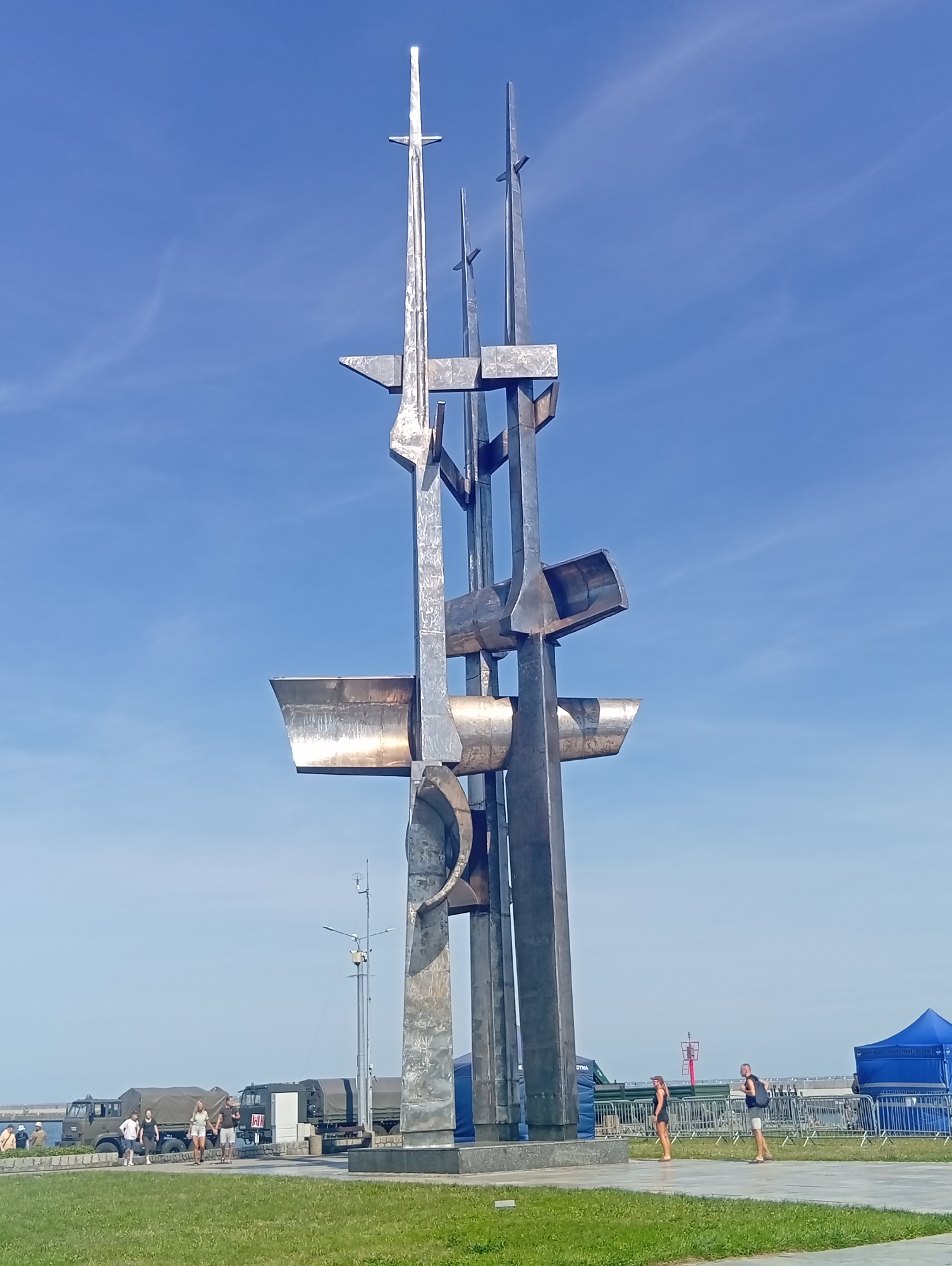 The image shows the Monument to the Fallen Shipbuilders in Gdynia, Poland. It is a tall, abstract sculpture made of metal, with various geometric shapes and vertical elements. The sculpture is situated outdoors, on a sunny day with a clear blue sky and some wispy clouds. In the background, there is a body of water, likely the Gdynia harbor, and various vehicles, including military trucks with canvas covers. Several people are visible in the foreground and midground, some walking and others standing. Two blue tents are set up on the right side of the image, and a red and white buoy or navigational marker is visible in the distance. The ground in the foreground is covered in green grass, with paved walkways and a low wall.