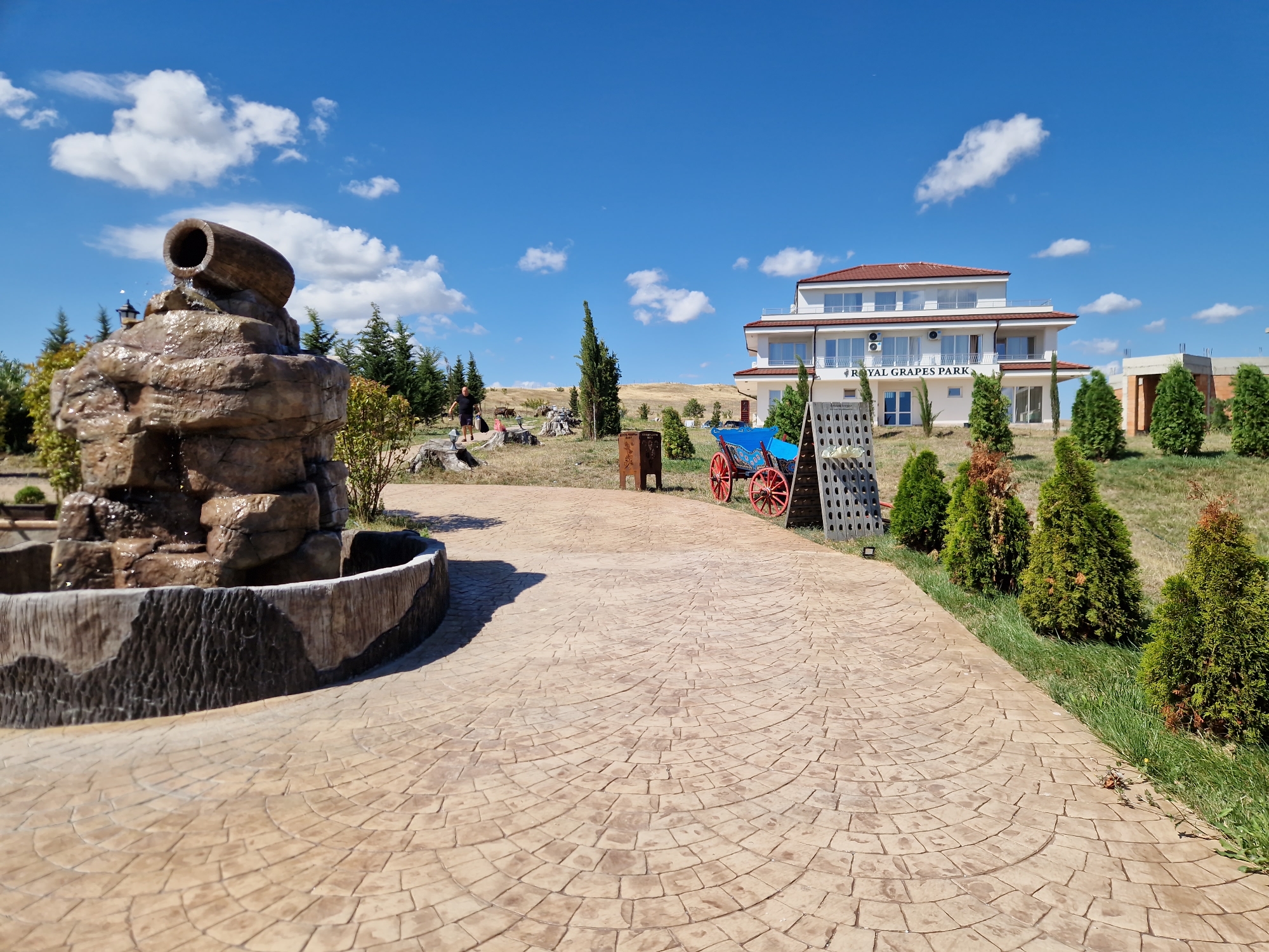 This image depicts the exterior of the "ROYAL GRAPES PARK" establishment in Tankovo, Bulgaria, under a clear blue sky with scattered white clouds. In the foreground, a decorative stamped concrete pathway curves to the right. To the left of the pathway is a rock-like fountain with a wooden log spout where water is visibly flowing out. Behind the fountain, a man in a dark t-shirt and shorts stands near some tree stumps. Further into the scene, a bright blue wagon with red wheels and a black frame is parked. A grey, lattice-like structure stands to the right of the wagon. Dominating the background is a multi-story white building with a red-tiled roof, displaying "ROYAL GRAPES PARK" signage. The building is surrounded by green cypress trees and rolling, dry grassy hills. The overall environment suggests a sunny, outdoor location, possibly a resort or entertainment venue.