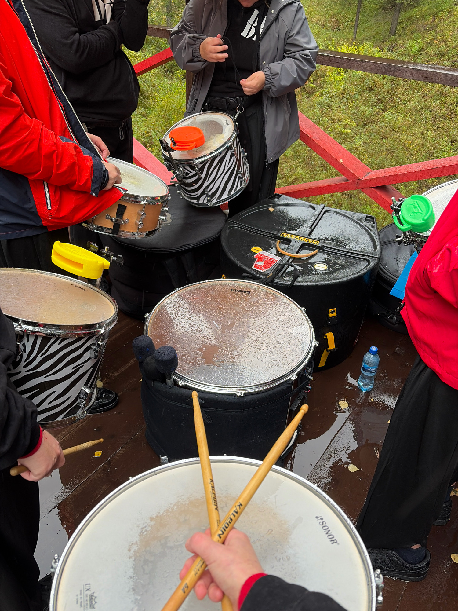 In Kukhtoozerskiy, Russia, a group of individuals are gathered on a wooden platform, likely outdoors, with a natural, vegetated background suggesting a park or forested area. The atmosphere appears wet, with raindrops visible on several drum surfaces and puddles on the wooden ground. Several drums are present, including a snare drum with a wooden body and metallic hardware, a tom-tom drum with a black and white zebra-like pattern, and larger bass drums, one of which is housed in a black hard case with yellow straps and branding. A smaller drum with an orange cymbal-like attachment is also visible. People are dressed in casual outerwear, including a red jacket, a grey hooded jacket, and black clothing. One person is holding drumsticks, poised as if to play a large drum in the foreground, which is also speckled with raindrops and has the "SONOR" logo on its head. Another drumstick rests on the wooden platform. A bottle of water is positioned near the drums. The overall scene depicts a drum ensemble preparing to perform or practice outdoors despite the rainy weather.