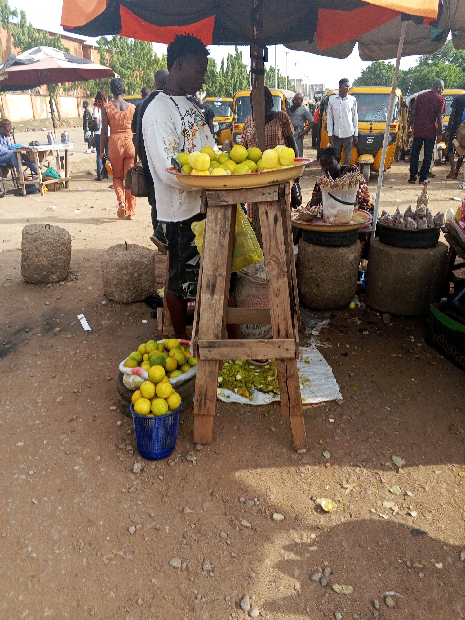 The image depicts an outdoor market scene in Abuja, Nigeria, characterized by informal trading setups and pedestrian traffic. A young man stands in the foreground, positioned behind a makeshift wooden table displaying a tray filled with yellow citrus fruits, likely lemons or limes. A blue plastic bucket filled with more citrus sits at the base of the table.
Behind him, multiple yellow auto rickshaws ("Keke Napep") are parked, suggesting this is a transportation hub or a busy commercial area. Several people are visible in the background, including pedestrians and individuals near other market stalls.
The setting includes rudimentary stone stools, possibly used as seating or display stands. Waste, including fruit peels, is scattered on the ground, indicating the practical, unrefined nature of the market environment. The scene is partially shaded by market umbrellas, providing relief from the sun.