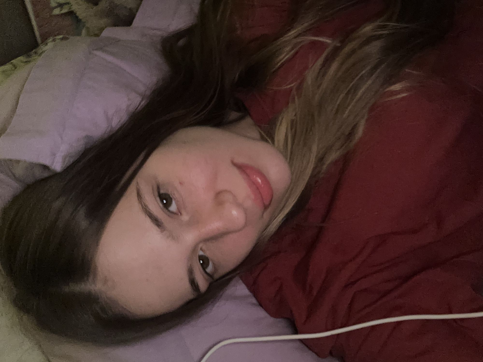 A young woman with long brown hair and light skin lies in bed, facing the camera at a slight angle. She is wearing a red shirt. Her eyes are open, and she has a subtle smile. The background shows a purple pillow and part of a bed. The image appears to be a selfie taken in a bedroom.