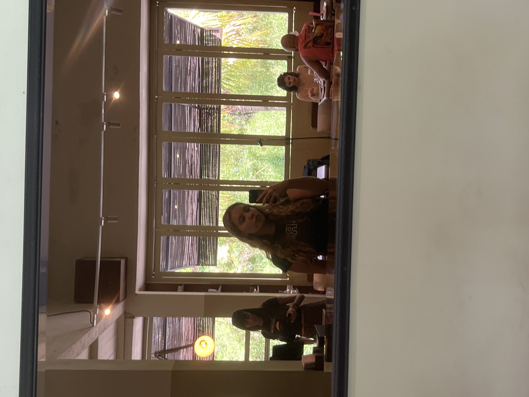 The image shows a reflection in a window or mirror. The reflection depicts the interior of a building with large windows offering a view of lush greenery. In the reflection, two people are visible taking a selfie with their phones. Additionally, the reflection shows two other people seated at a table. The location is possibly in North Kuta, Indonesia, inferred from the vegetation and architectural style seen through the windows. The interior appears to be a restaurant or open-air lounge area with interior lighting.
