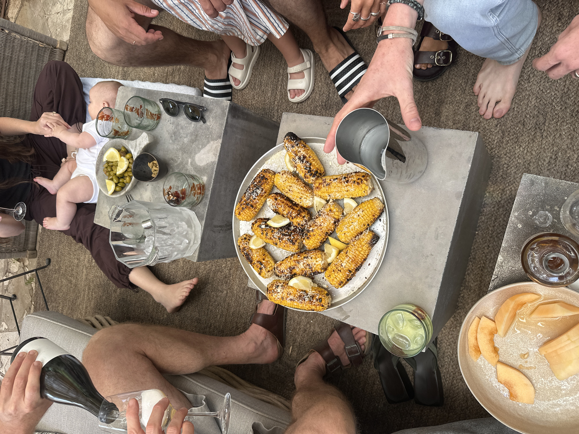 The image shows an outdoor dining scene. Several people are seated on the ground and chairs surrounding two low square tables. A baby wearing a white outfit is being held in someone's lap.
On one of the tables are a plate of green olives and lemon slices, sunglasses, and several glasses. A person pours liquid from a pitcher onto a plate of grilled corn on the cob that is on the other table. The second table also holds a glass of a light-colored liquid with ice and a plate of sliced cantaloupe.
The floor is covered in a neutral-toned rug. The scene implies a casual gathering, possibly in the city of Tel-Aviv, Israel, but no specific landmarks or characteristics confirm this location.