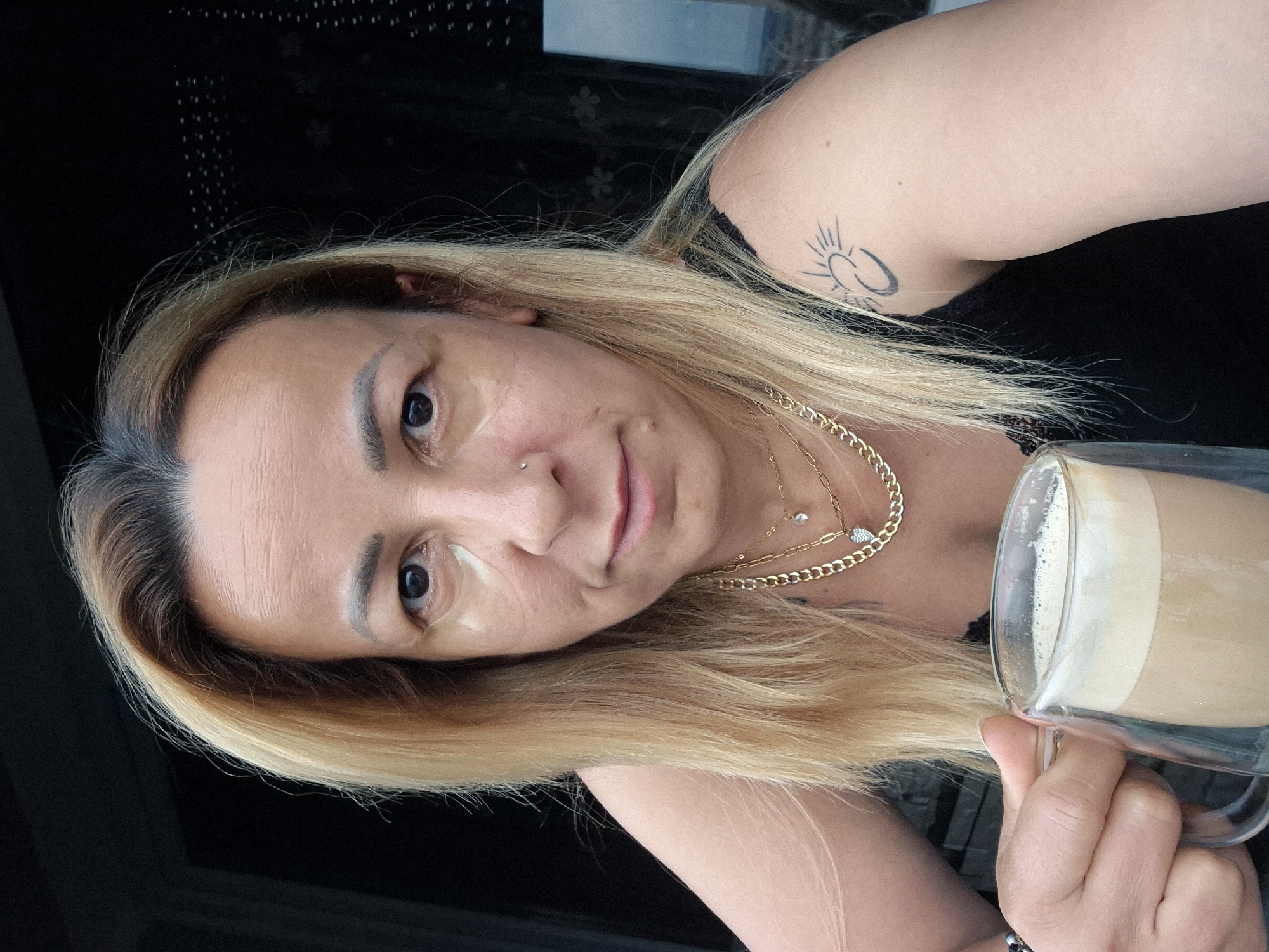 A woman with blonde hair and tanned skin is holding a clear glass mug filled with a foamy, light brown beverage. She is looking directly at the camera with a neutral expression. She is wearing a black tank top and several gold necklaces, one of which has a small heart pendant. There is a tattoo of a crescent moon and sun on her upper right arm. The background is dark and indistinct, suggesting an indoor or shaded outdoor setting. The image appears to be a selfie.