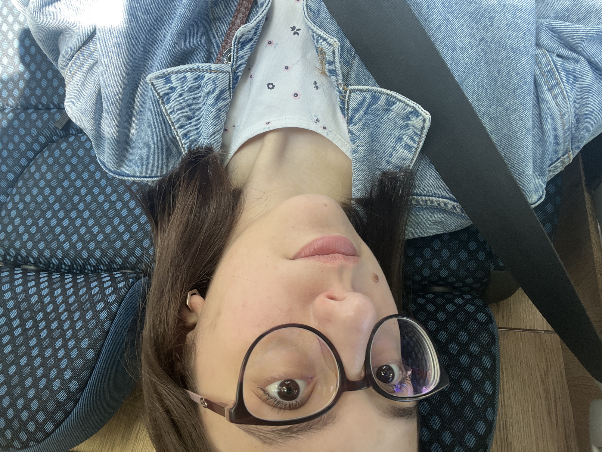 A woman wearing glasses, a denim jacket, and a white floral-patterned shirt is secured in a seat with a black seatbelt. The seat has a dark blue fabric cover with a small diamond pattern. Her hair is dark brown. The location of the photo is unknown and cannot be determined based on the image. The statement "the location in the city of Riahovtsite, Bulgaria" cannot be verified based on the image.