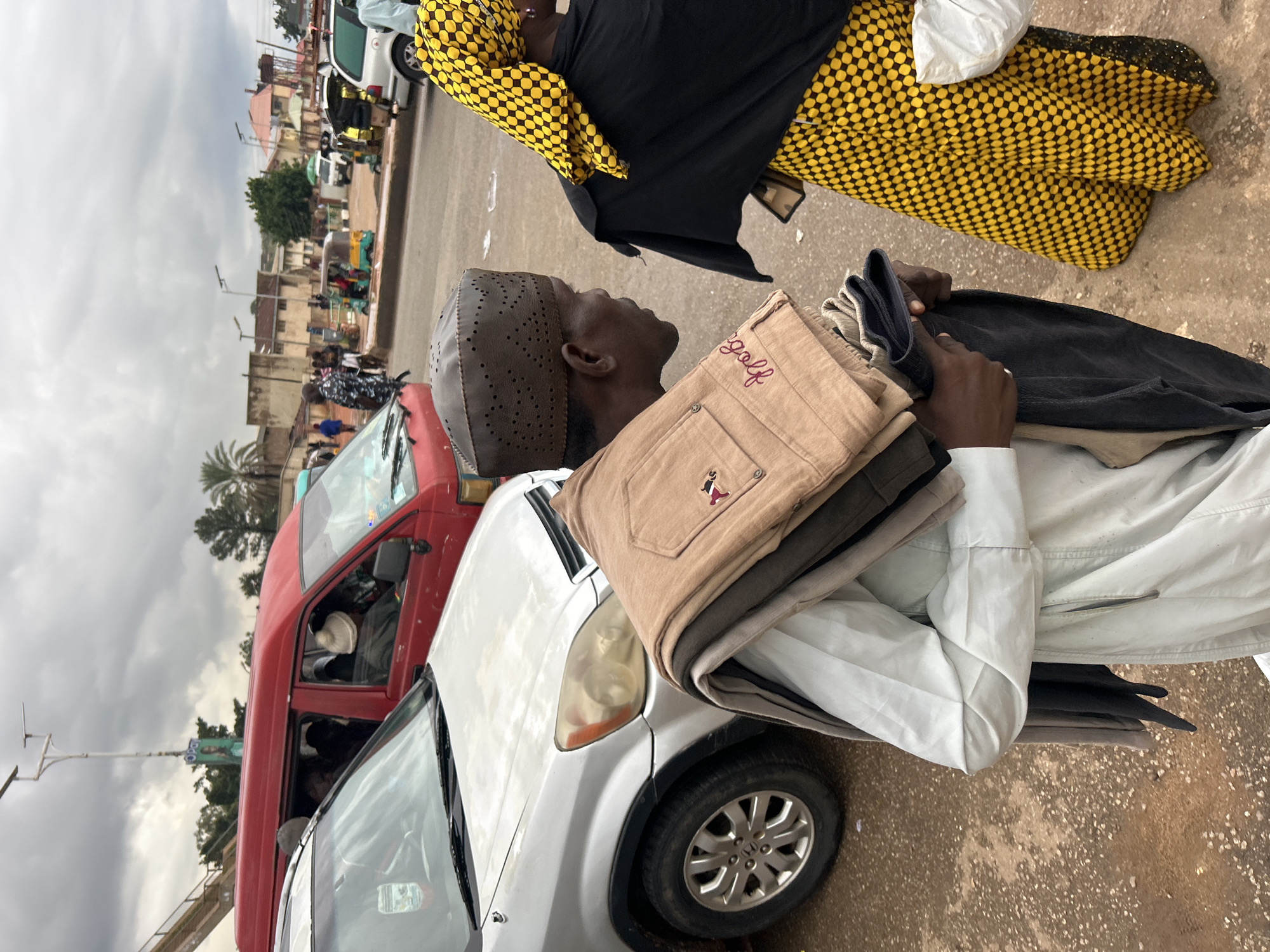 In Jos, Nigeria, on a cloudy day, a man in a dark perforated cap and a light-colored tunic is walking, carrying folded clothes. The clothes, primarily in tan and dark colors, are draped over his left arm and shoulder. He is walking beside a silver SUV. In the background, a red minibus, other vehicles, and buildings suggest a bustling urban environment. Further back, a woman in a black and yellow checkered headscarf and a yellow patterned dress is visible, her back turned to the camera. The scene captures a moment in the daily life of Jos.