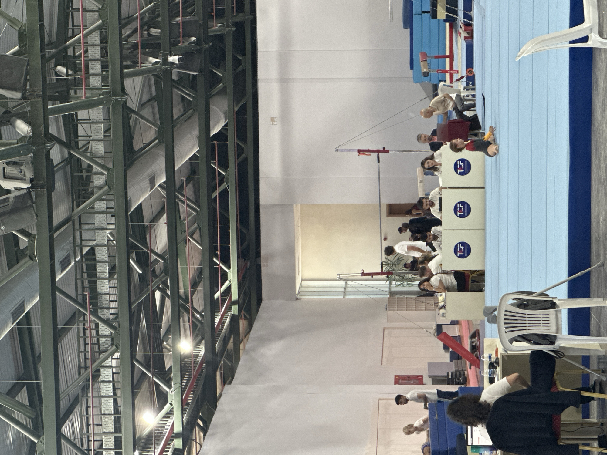This image depicts a gymnastics competition or training session within a large indoor facility in Sancaktepe, Turkey. The scene is characterized by a high ceiling with exposed industrial lighting and ductwork. In the foreground, a blue gymnastics mat covers a significant portion of the floor. A young girl in a leotard is sitting on the mat, exhibiting a straddle split.
Several people are visible in the middle ground, positioned behind a series of white partitions with "TCF" logos. These individuals appear to be officials or judges, some interacting with laptops or other equipment. A few athletes, possibly waiting for their turn or observing, are also present in this area. Further back, individuals are seen near stacked blue mats, suggesting equipment storage or preparation.
The background shows more of the expansive gymnasium, with stark white walls and the continuation of the industrial ceiling structure. The lighting in the facility appears to be a mix of overhead fluorescent and spotlights. The overall environment suggests a professional or semi-professional sporting venue.