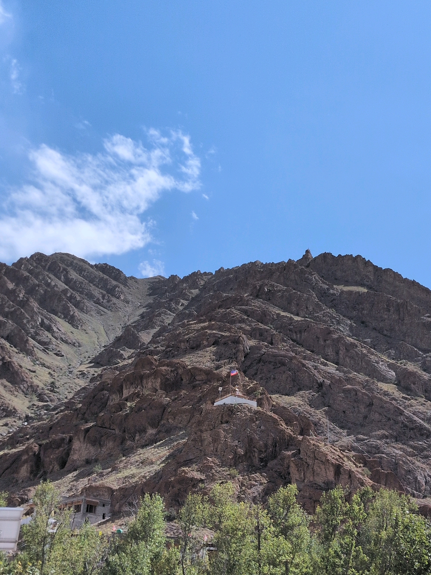 The image shows a vast, rugged mountain landscape under a bright blue sky with scattered white clouds. The mountains are steep and rocky, with visible geological strata and scree slopes. In the mid-ground, nestled on a rocky outcrop, is a small, white structure topped with flags. A red and blue flag flies prominently from a pole. Below the outcrop, a cluster of buildings with traditional architecture, featuring stone construction and wooden elements, is visible amidst lush green trees. The scene is devoid of people. The overall impression is one of natural grandeur and a remote, mountainous settlement, likely in or near Hemis, India, known for its monastery and high-altitude terrain.