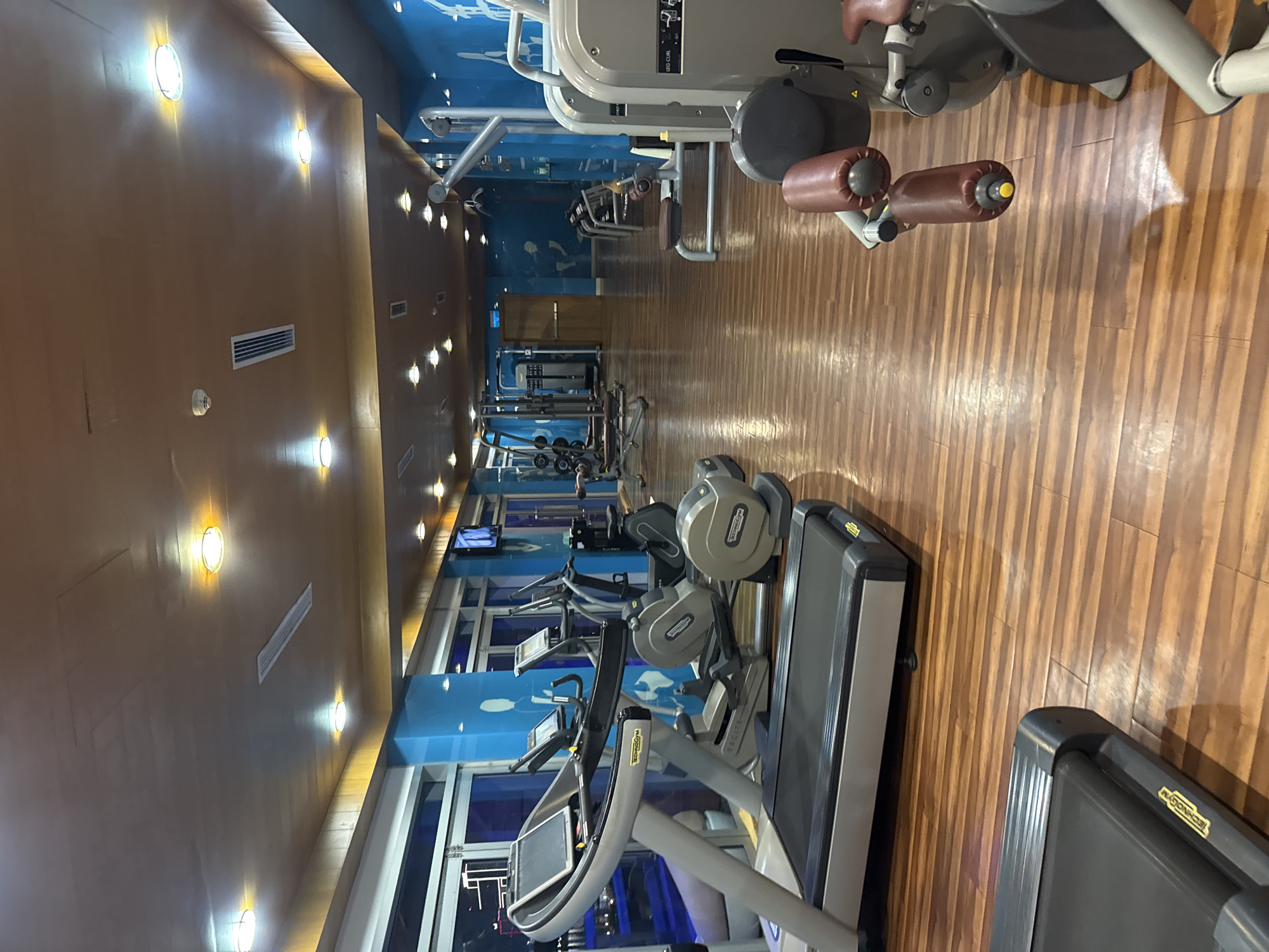 The image depicts the interior of a gym in Đà Nẵng, Vietnam. The gym features wood flooring and a light brown ceiling with recessed lighting. The walls are blue with ocean-themed decor. Exercise equipment, including treadmills and weight machines, is arranged along the length of the gym. No people are visible in the image.