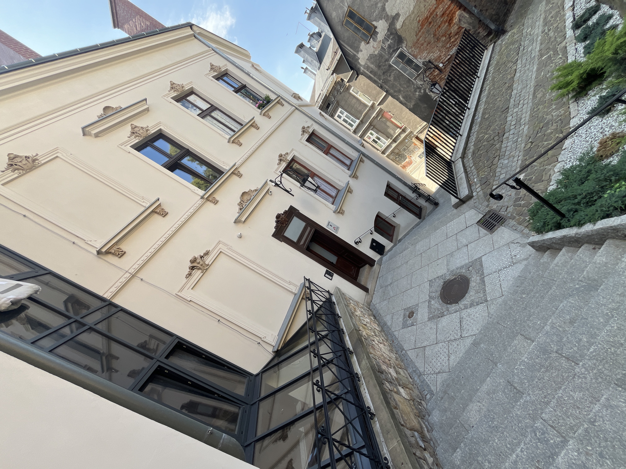 An outdoor, eye-level, full shot captures a narrow, descending stone staircase in an urban setting. The staircase is flanked by stone walls on the left and a decorative metal railing and planted shrubbery on the right. The stairs are made of large, light-grey granite slabs. The stone wall on the left is constructed from irregular, tan and grey stones. The area at the top of the stairs features a paved walkway with square, grey tiles and a circular metal manhole cover.
The buildings lining the staircase vary in style and condition. On the left, a cream-colored building with classical architectural details, including ornate decorative elements above the windows and along the upper facade, is visible. This building features large, dark-framed windows, one of which is a modern glass-paneled structure with a dark metal frame and railing. A dark wooden door with a glass pane is also present.
To the right of the cream-colored building, a series of other buildings are visible. One is a grey building with visible brickwork and some stucco damage, showing a rough, weathered facade. This building has multiple windows, some with visible panes and frames. Further down the staircase, another building with lighter colored walls and smaller windows is visible. A dark metal railing runs along a stone-lined walkway partway down the stairs. The sky above is a clear, bright blue with some wispy clouds. The image is taken at an upward angle, giving a sense of depth and perspective.