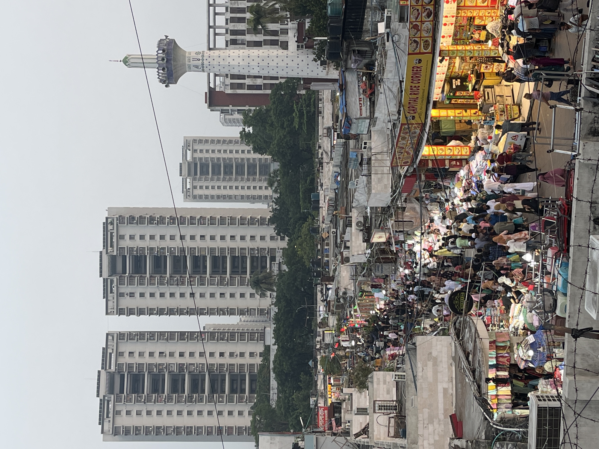 The image depicts a crowded market street in Dhaka, Bangladesh, as indicated by the presence of the Baitul Mukarram National Mosque visible in the background. The scene is bustling with numerous people shopping and walking through the narrow street lined with shops and stalls. Several tall apartment buildings can be seen in the distance behind the market area. The lighting suggests it may be dusk or early evening. The street is filled with various goods and products displayed for sale. Overhanging wires and signs are visible overhead, adding to the congested feel of the scene.