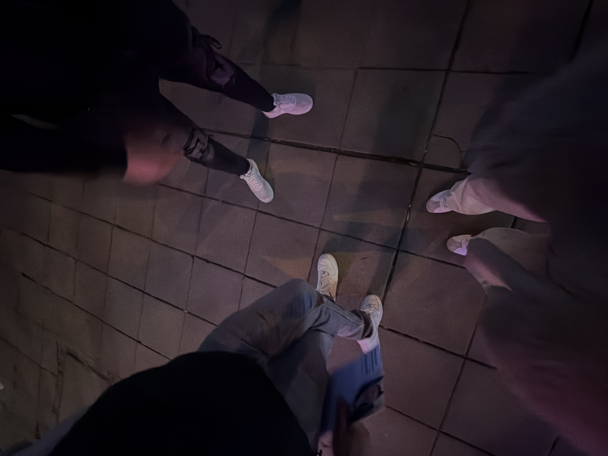 The image depicts a low-angle view of the lower bodies of at least three individuals standing on a tiled pavement at night in Sofia, Bulgaria. The ground consists of dark-colored square or rectangular paving stones with visible grout lines. Dim artificial lighting casts a reddish-purple hue across parts of the scene.
From left to right:
1. One individual wears light blue jeans and white athletic sneakers, with a dark upper garment partially visible.
2. Another individual, positioned at the bottom center of the frame, wears light-colored trousers and white athletic sneakers.
3. A third individual, positioned on the right, wears dark-colored pants with lighter patterns or distressed areas on the knees, and white athletic sneakers.
All individuals appear to be standing or in motion on the pavement. No specific interactions are discernible.