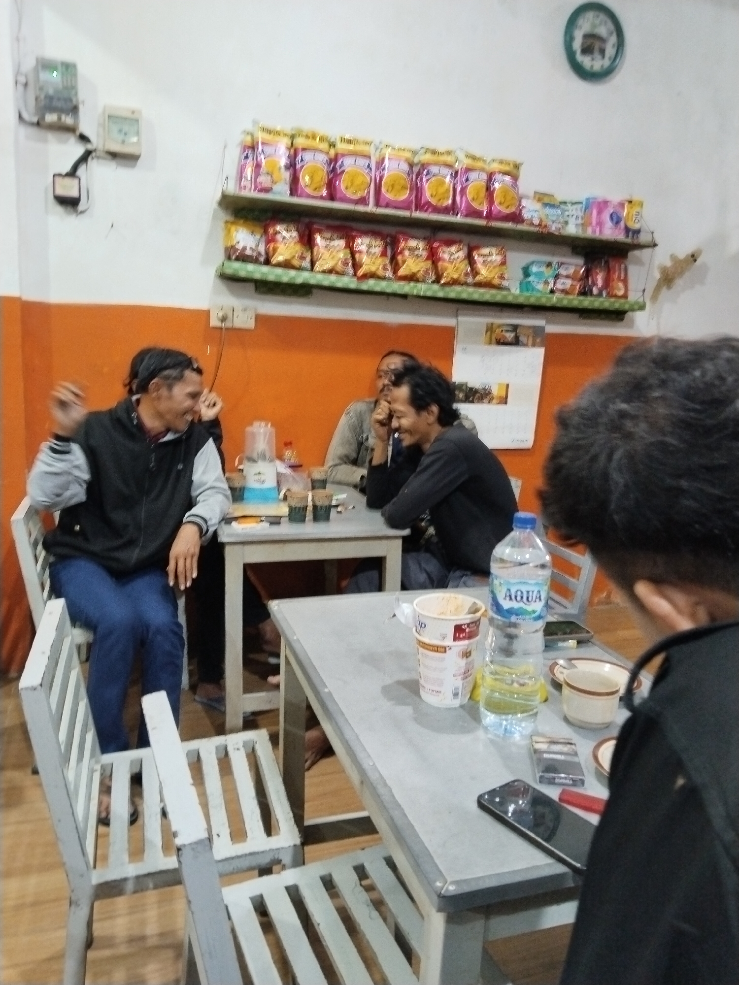The image depicts an indoor scene, likely a small eatery or shop, in Prigi, Indonesia. Several individuals are seated around tables, engaged in conversation and consumption. On the left, a person wearing a black and grey jacket and blue jeans is seated on a white slatted chair, gesturing with their right hand. They appear to be laughing or speaking animatedly. A blender and cups are visible on a table in front of them.
Further into the scene, two other individuals are seated at a table. One person, with dark, curly hair, is wearing a dark long-sleeved shirt and appears to be looking towards the left, also seemingly engaged in conversation. Another person is partially visible in the foreground, with their back to the camera, their dark, curly hair prominent.
The environment features orange-painted walls and white shelving units displaying various bagged snack items. A clock with a picture of the Kaaba is mounted on the wall. Electrical outlets and wiring are visible on the wall. A bottle of Aqua brand water, a cup of what appears to be instant noodles, a pack of cigarettes, a phone, and a lighter are on a table in the foreground. The tables are made of a light-colored material, and white slatted chairs are utilized. The lighting suggests an indoor setting, possibly during evening hours.
