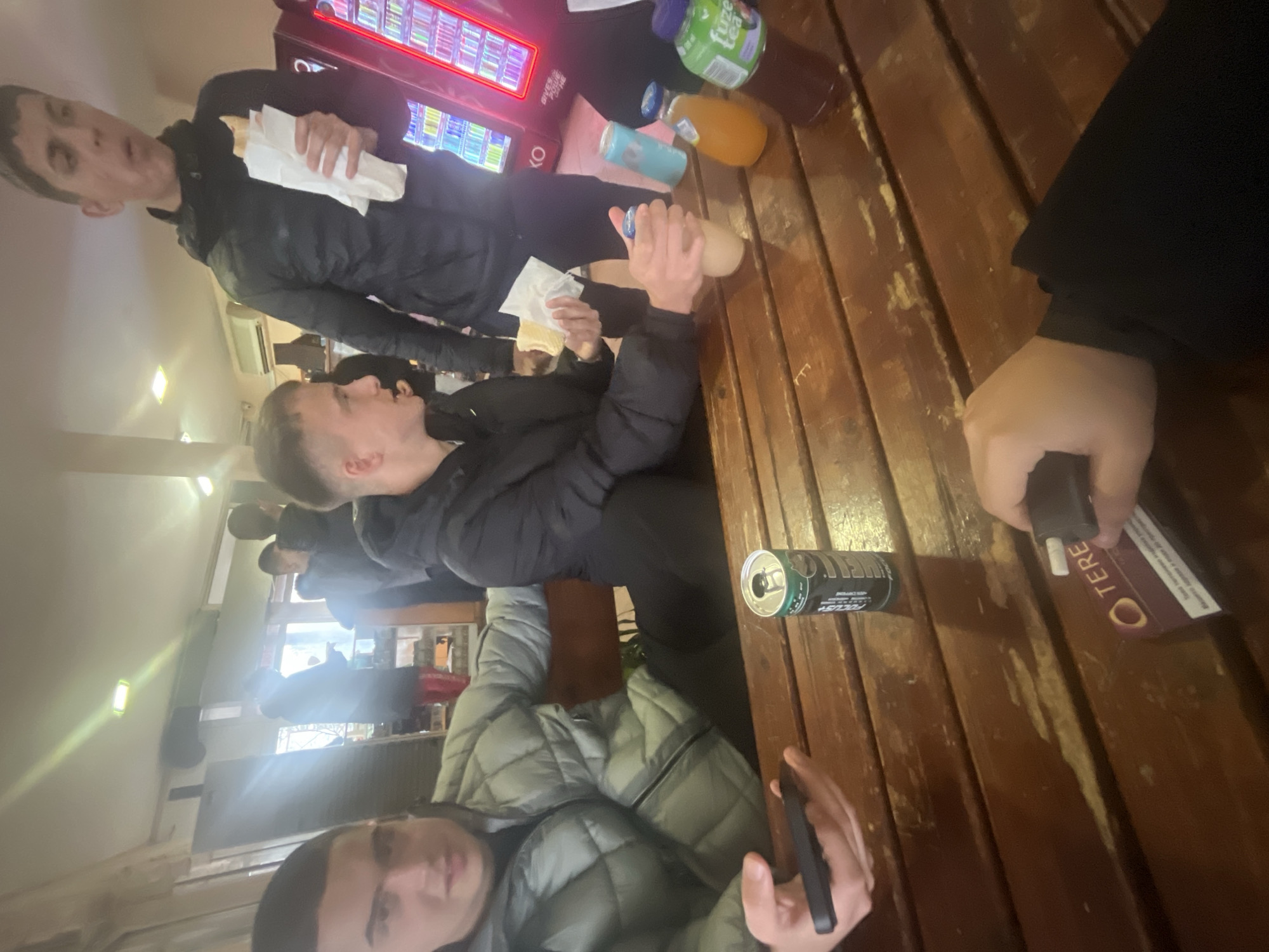 A medium shot depicts a group of at least five people seated around a wooden table. The setting appears to be a casual eating area, possibly in Asenovgrad, Bulgaria, given the context. The individuals are dressed in casual attire, including puffer jackets, and are interacting with objects on the table such as food, beverages, and mobile phones. A vending machine is visible in the background.