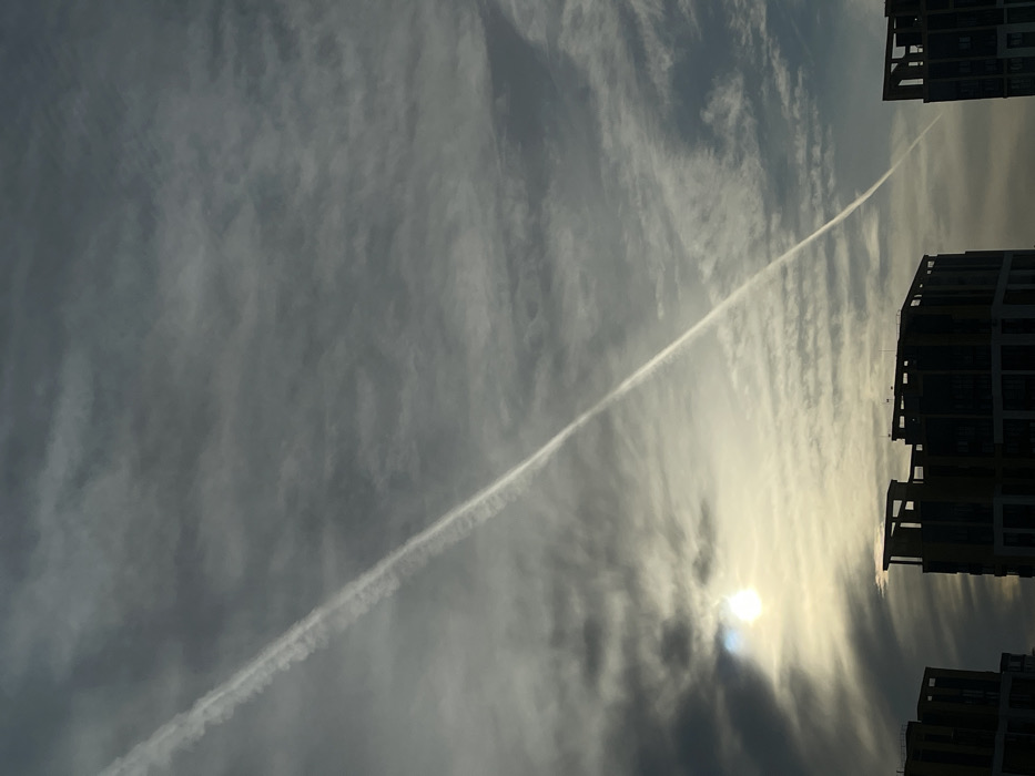 The scene, located in Izhevsk, Russia, depicts an urban sky dominated by a prominent white airplane contrail stretching diagonally from the upper left to the lower right. The sky is primarily overcast with a mixture of dark and lighter gray clouds, exhibiting varying textures. The sun, appearing as a bright, diffused white and yellow glow, is partially obscured by clouds in the lower-left quadrant. Silhouetted against the bright horizon and occupying the bottom portion of the frame are the dark upper sections of at least three modern, multi-story buildings, characterized by flat roofs and dark window patterns. No individuals or active interactions are visible.