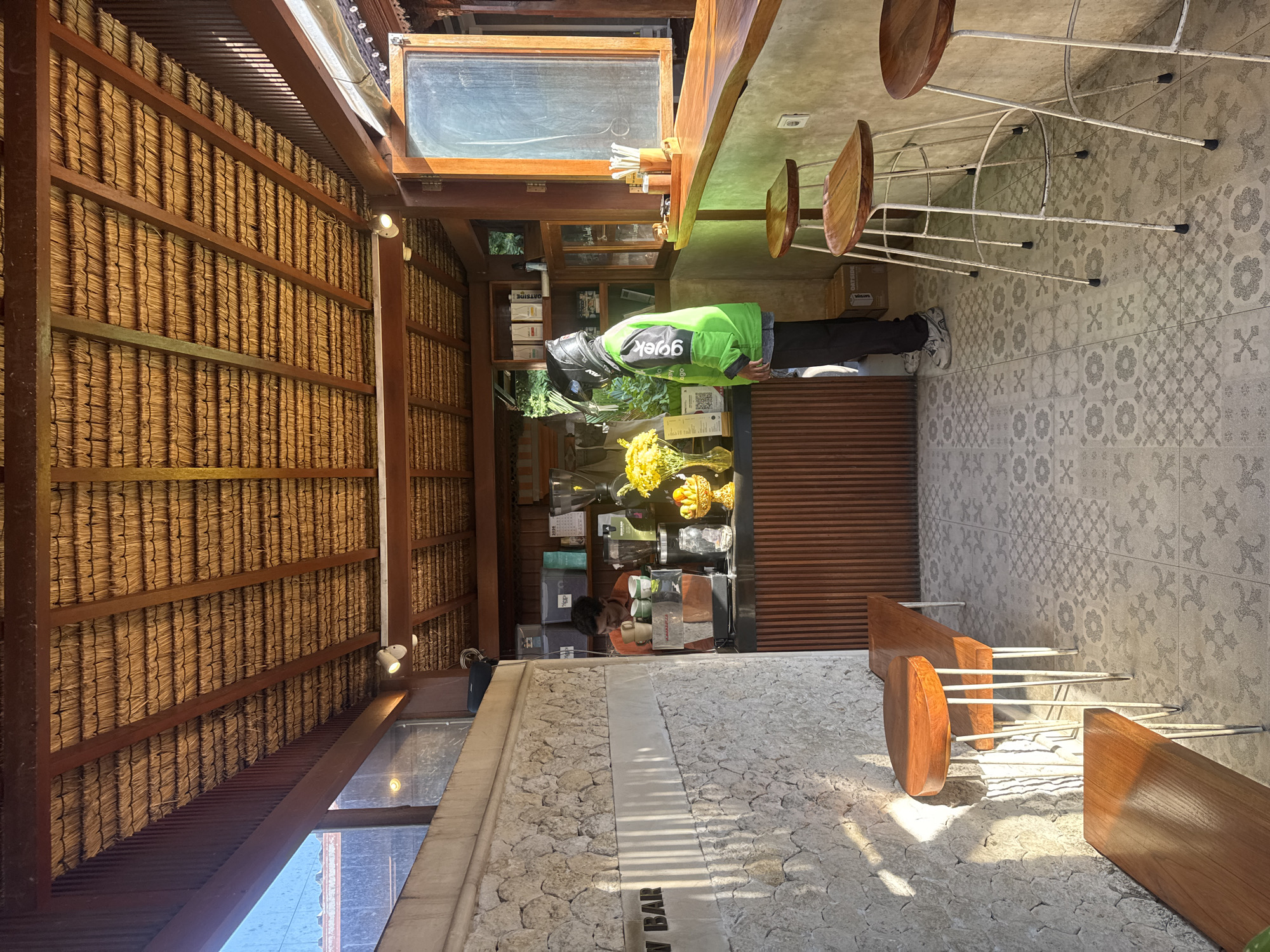 The image depicts a coffee shop with an outdoor seating area. In the foreground, to the left, are two wooden benches with metal legs. The walls are constructed from textured stone with vertical wooden accents. The floor is tiled with a repeating geometric pattern. In the middle ground, a barista in an orange shirt is seen behind a counter, preparing drinks. To the right, a delivery driver wearing a green Gojek jacket and a helmet stands at the counter. The counter is a live-edge wooden slab with several bar stools in front of it. The ceiling is thatched with exposed wooden beams. The background reveals shelves stocked with coffee-related items and a window opening to the outside. The scene is lit by natural sunlight.