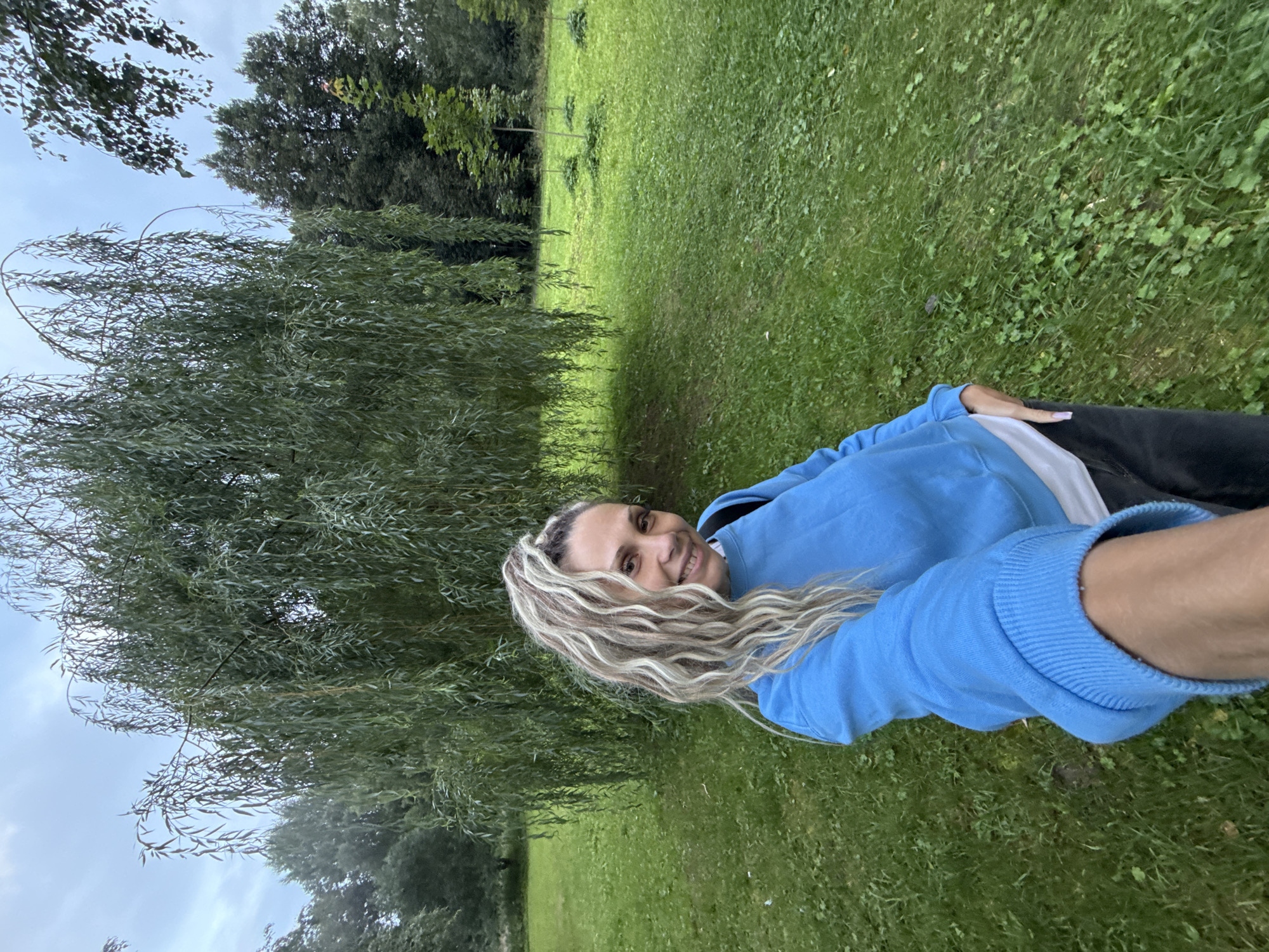 The image shows a woman with long, wavy blonde and brown hair styled in a high ponytail, wearing a bright blue sweatshirt over a white t-shirt and dark pants. She is smiling and holding the camera, taking a selfie. She is standing in a grassy park with a large weeping willow tree behind her. The background shows more trees and a cloudy sky. The location is identified as Rusanovo, Russia.